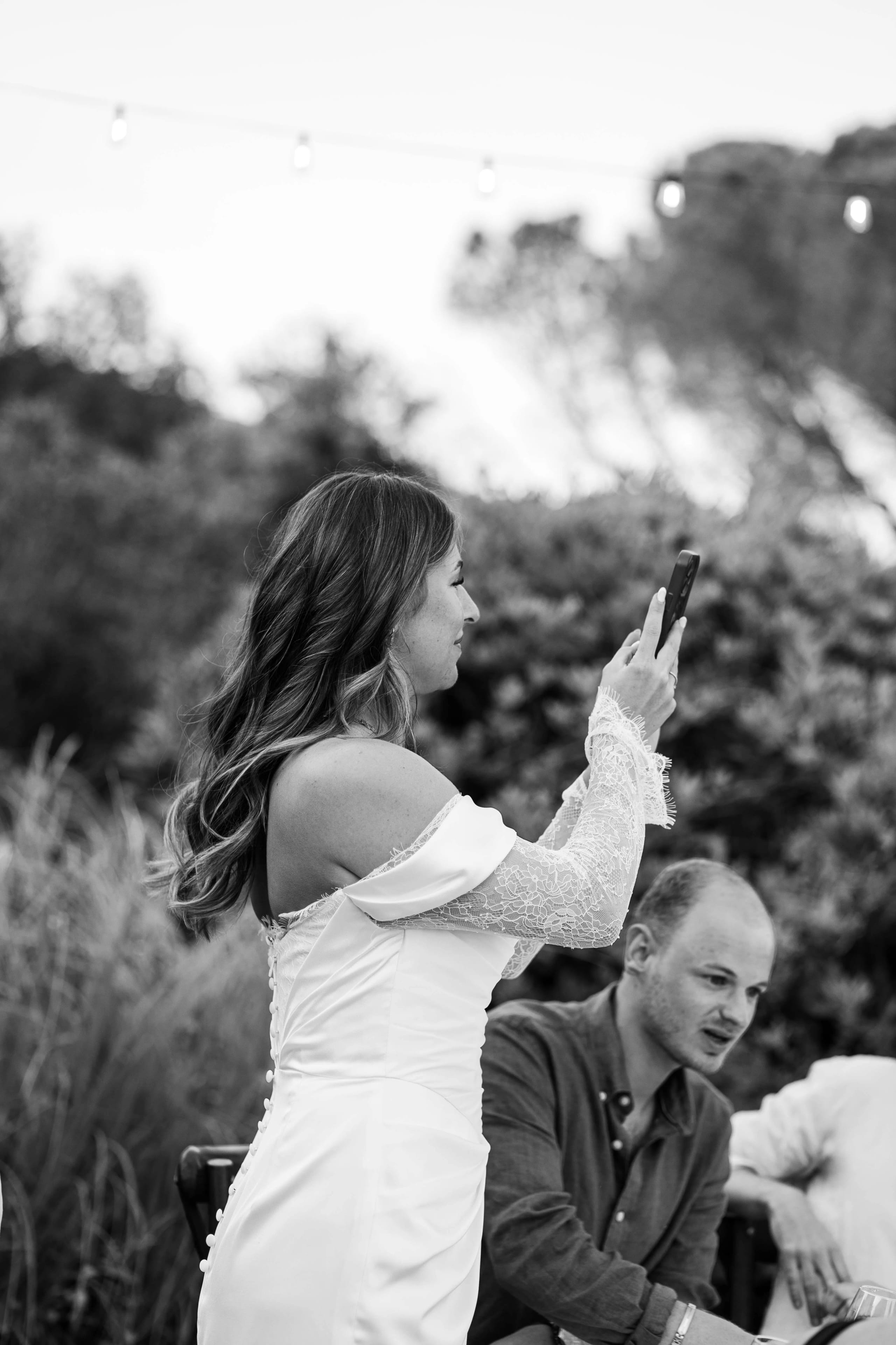 Een vrouw in een elegante witte jurk maakt een foto met haar smartphone terwijl ze in een groene, ontspannen omgeving staat, met een man in een casual outfit die naast haar zit.