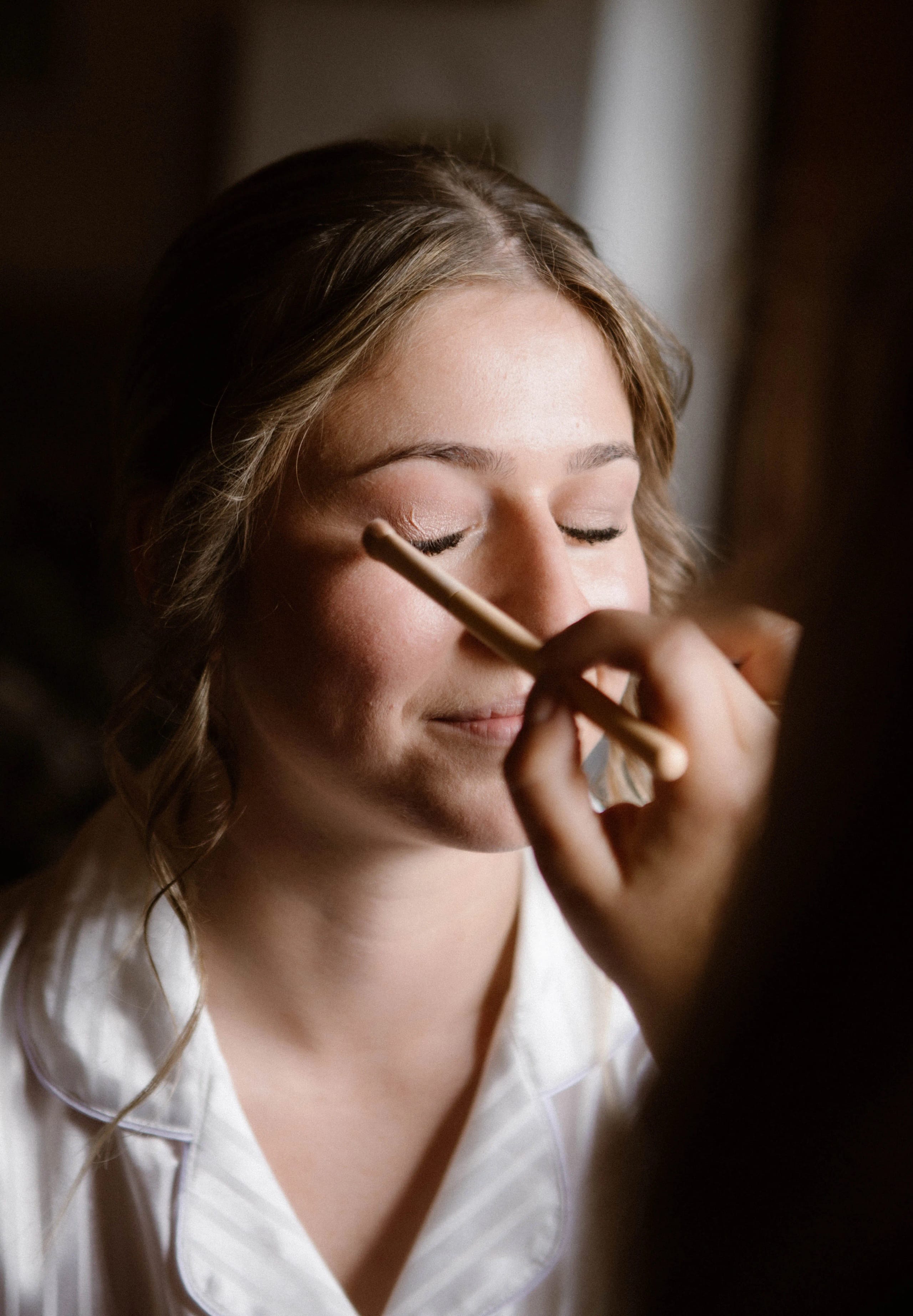 Een jonge vrouw met een zachte glimlach krijgt make-up aangebracht terwijl ze ontspannen haar ogen sluit, omringd door een warme, sfeervolle achtergrond.