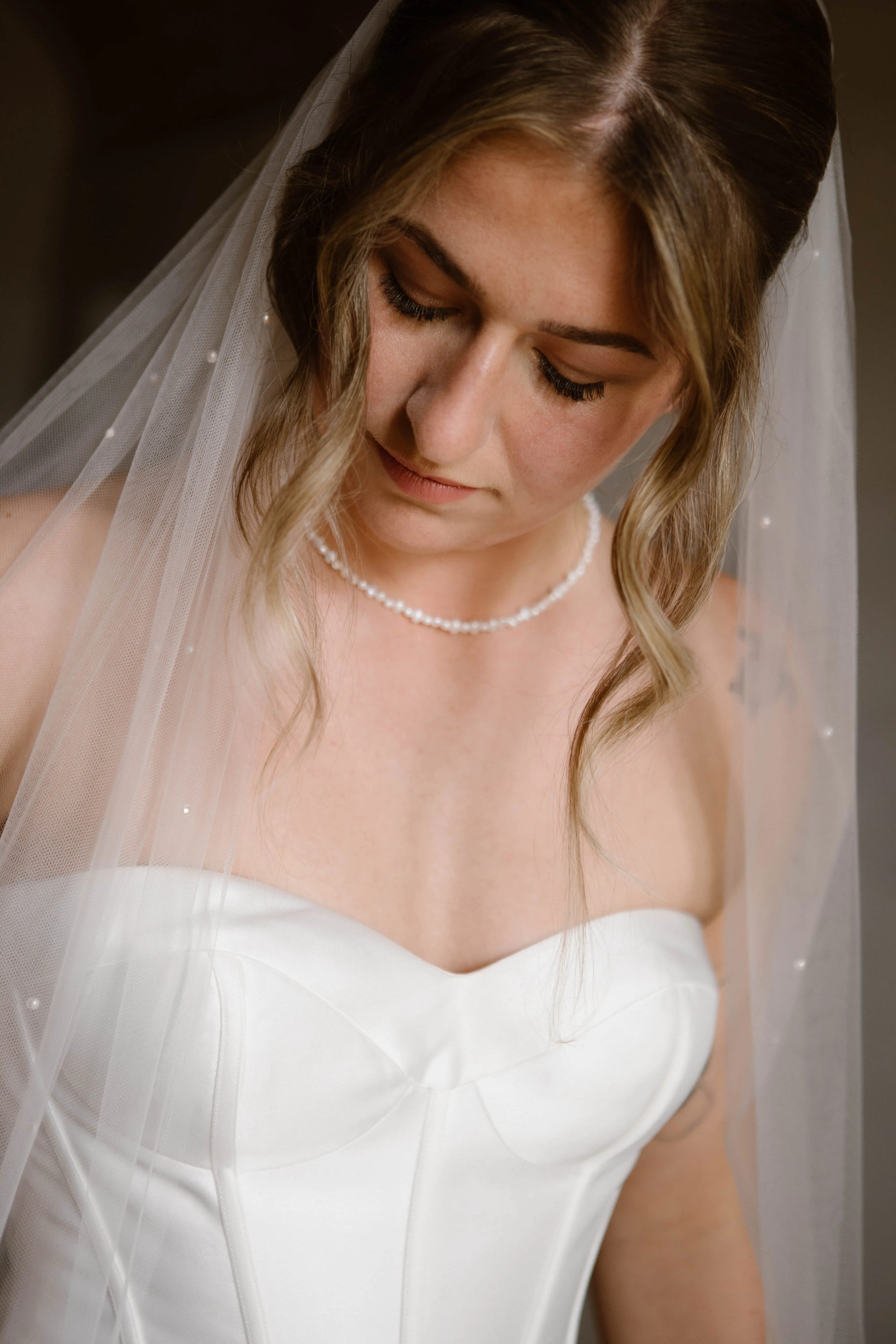 Een bruid in een schitterende strapless trouwjurk met een lange, delicate sluier, die voorzichtig naar beneden kijkt en haar accessoire aanraakt, terwijl ze zich voorbereidt op haar speciale dag.