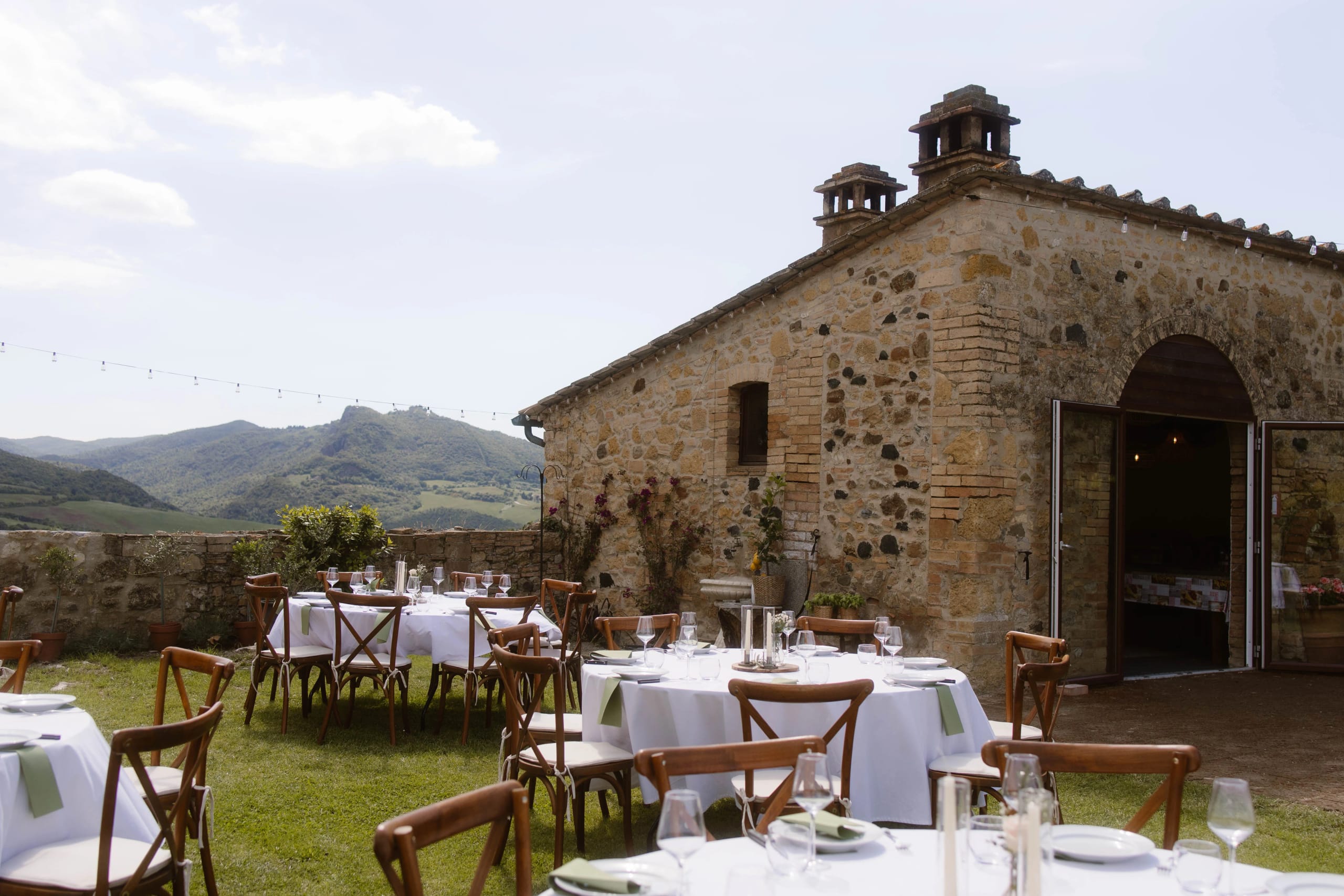Een charmant buitenrestaurant met elegant gedekte tafels en houten stoelen, gelegen in een schilderachtige landschap met heuvels en een vintage stenen gebouw op de achtergrond.