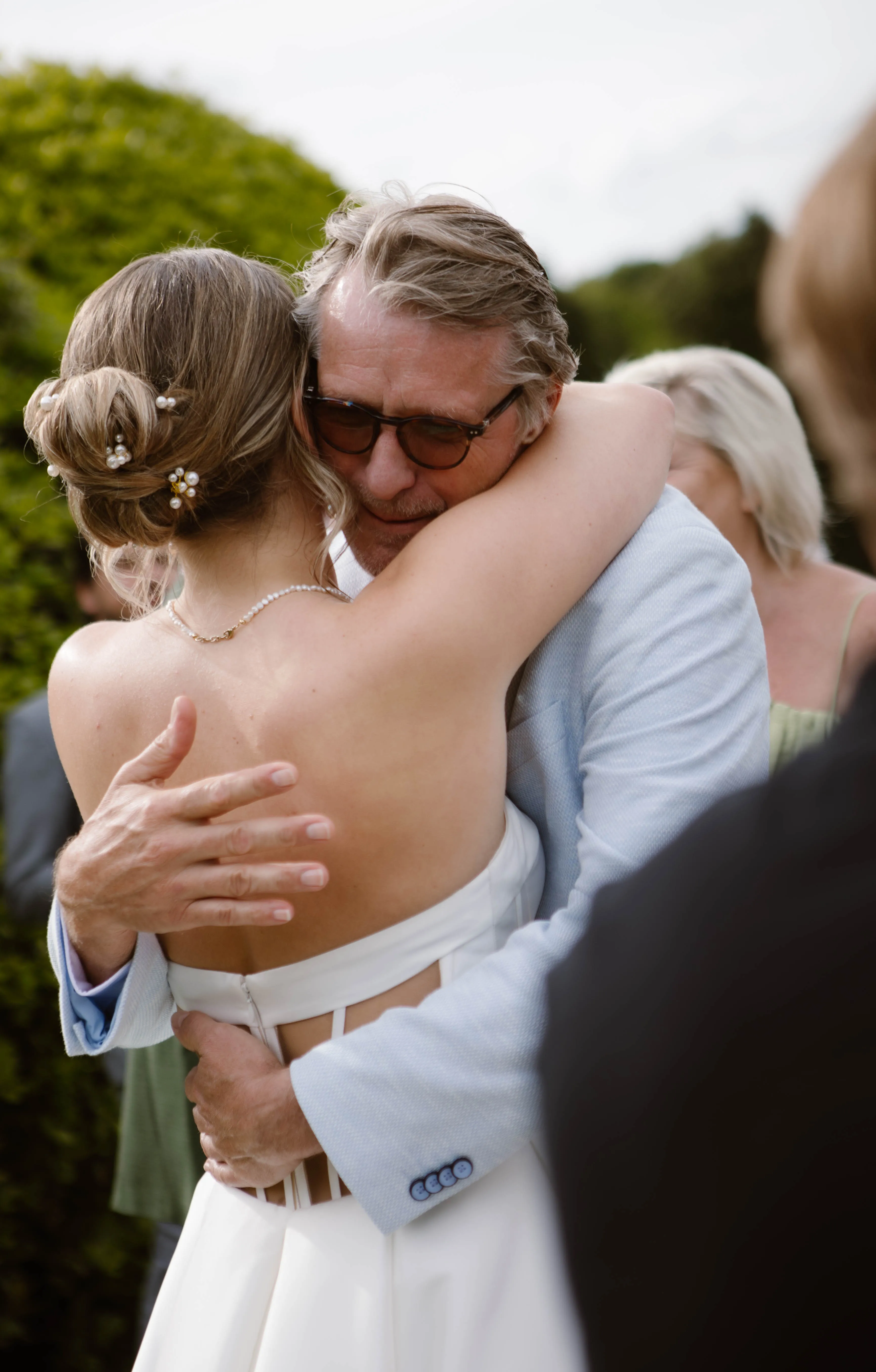Een emotionele omhelzing tussen een bruidegom en zijn dochter in een prachtige tuin, met een achtergrond van weelderig groen en gasten die genieten van het huwelijk. De bruid draagt een elegante witte jurk en haar haar is versierd met bloemetjes, terwijl de vader een lichtblauwe blazer aanheeft.