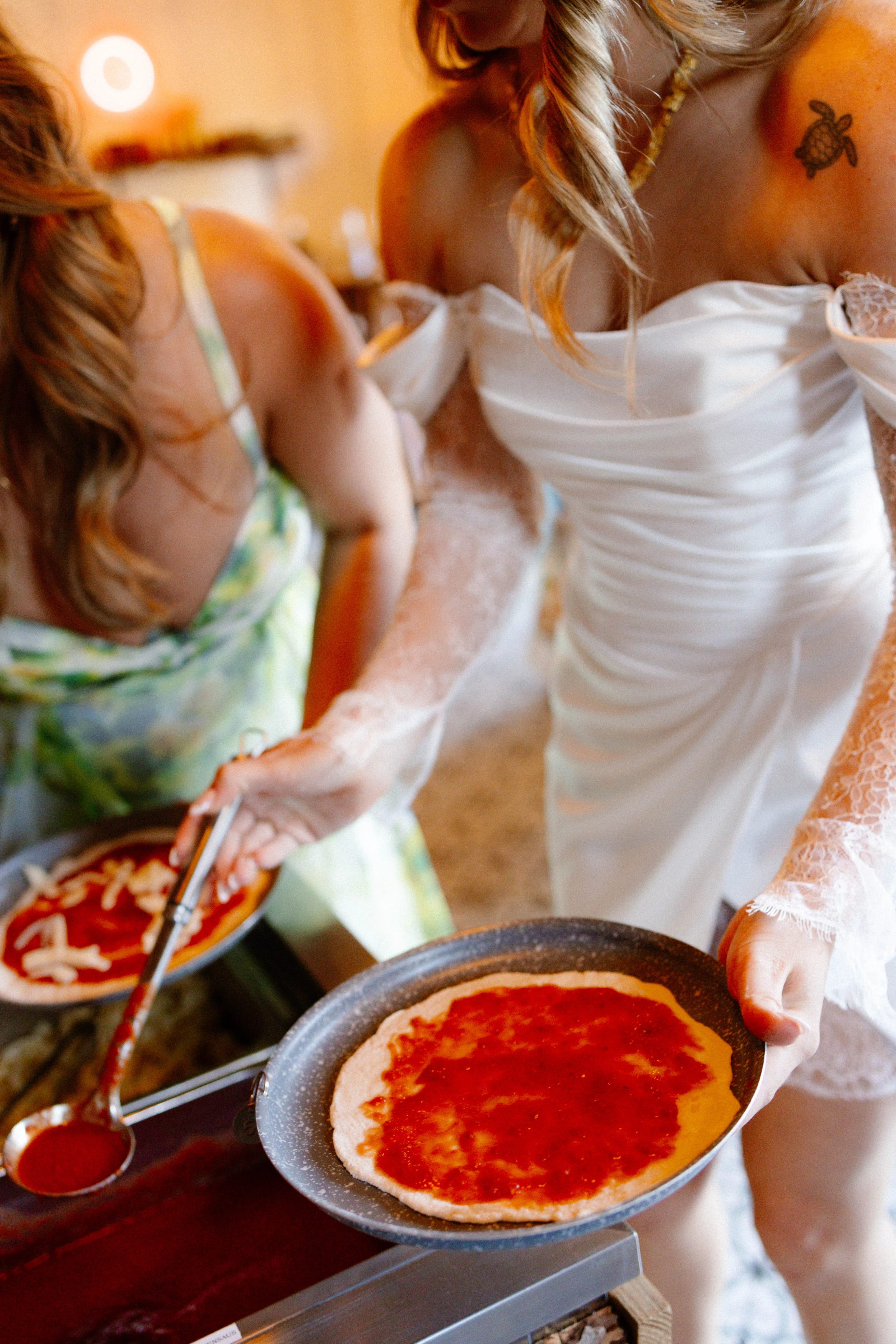 Een bruid in een feestelijke witte jurk druppelt tomatensaus op een pizzabodem terwijl een andere vrouw, gekleed in een kleurrijke jurk, dichtbij staat. De sfeer is informele en vrolijke kookactiviteit tijdens een bruiloft.