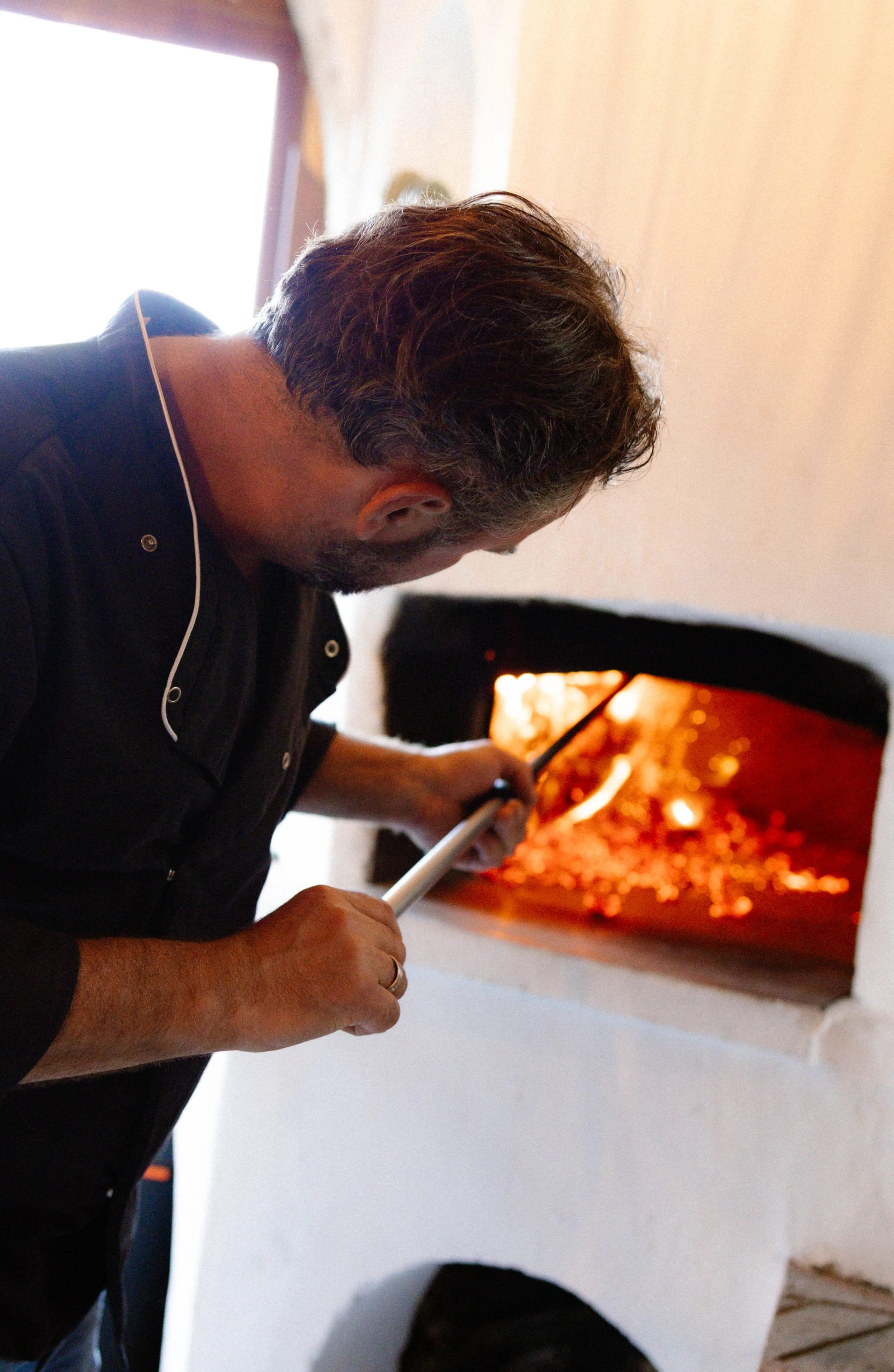 Een man in een zwart overhemd staat gebogen over een traditionele houtgestookte oven, terwijl hij met een lange stok de vlammen en het vuur in de oven aanstuurt. De warme gloed van het vuur geeft een gezellige sfeer aan de ruimte.