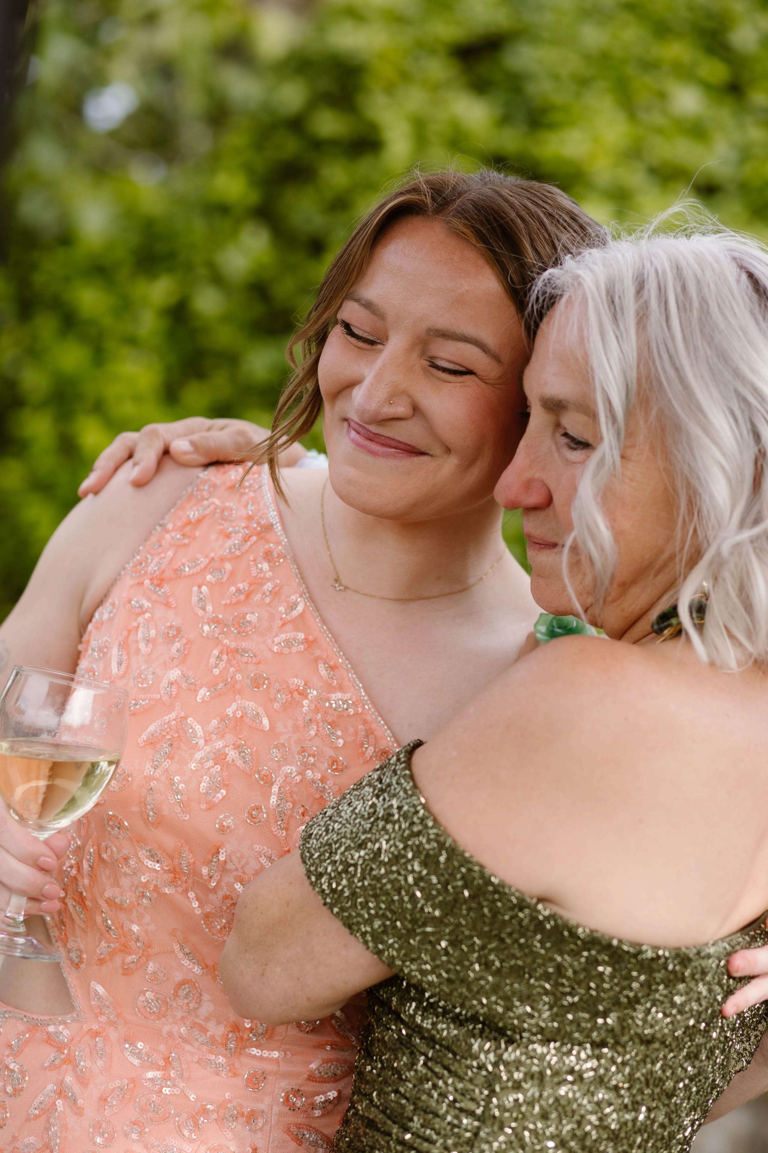 Twee vrouwen omhelzen elkaar blij en stralend, terwijl de vrouw links, gekleed in een glinsterende perzikkleurige jurk en met een glas witte wijn in de hand, de vreugdevolle verbinding viert met de vrouw rechts in een glitterende groene jurk. De achtergrond bestaat uit weelderig groen, wat bijdraagt aan de feestelijke sfeer.