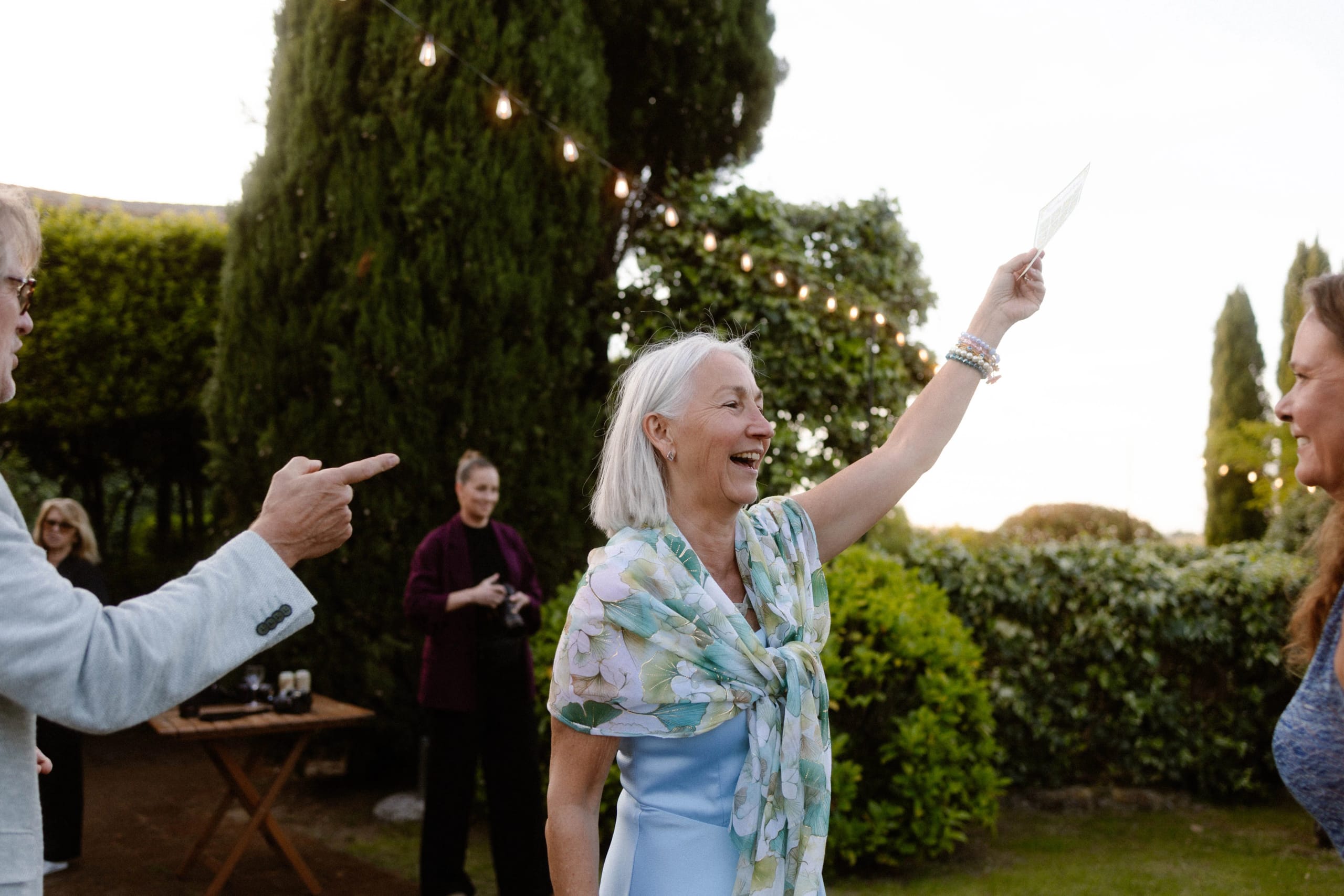 Een vrouw met een stralende lach steekt een kaart omhoog terwijl ze in een tuin staat, omringd door groene struiken en sfeervolle verlichting, terwijl een man naar haar wijst.