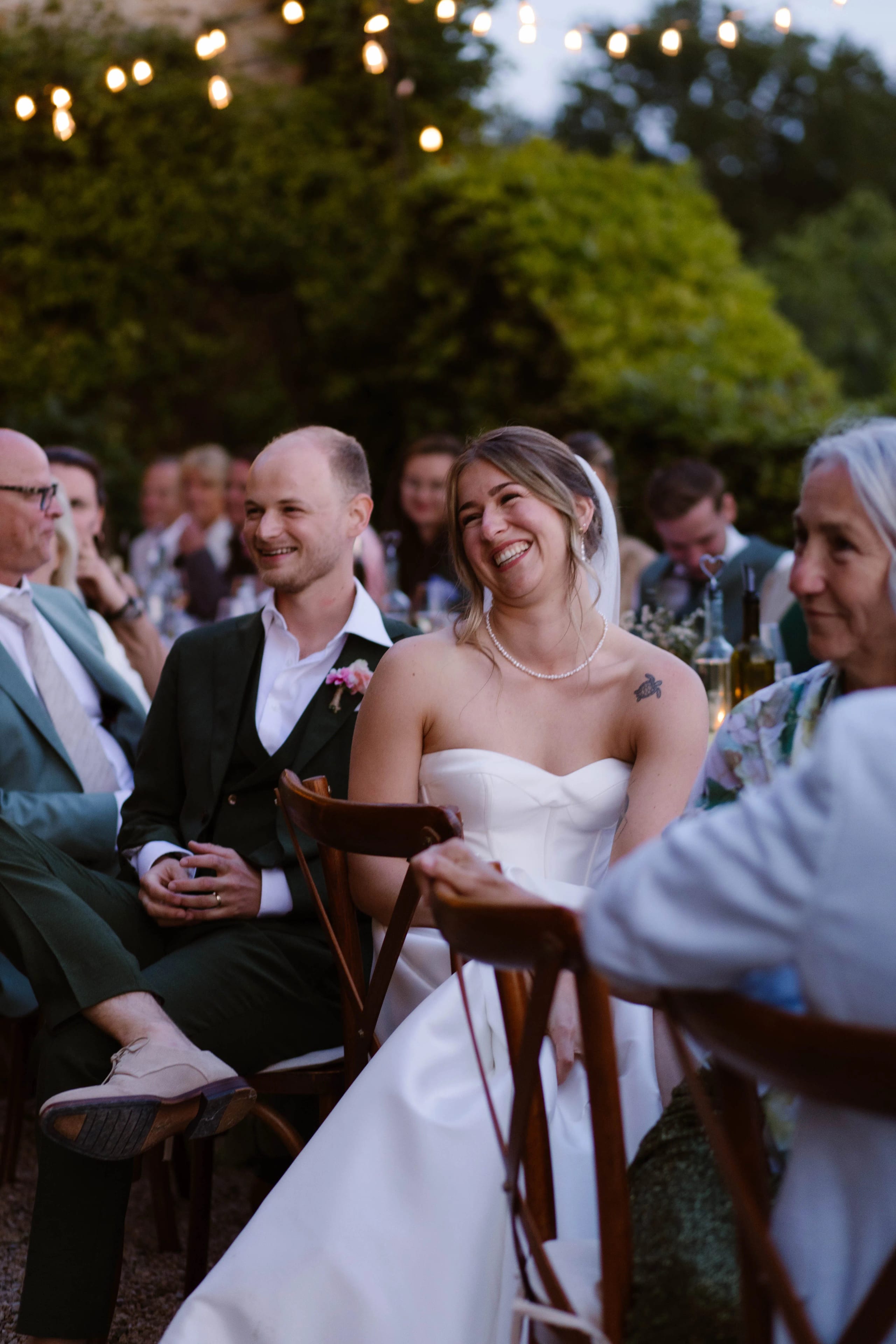 Een blije bruid in een strapless witte jurk lacht terwijl ze praat met de man naast haar, terwijl gasten aan een prachtig verlichte trouwtafel genieten van de sfeer van het buitenfeest.