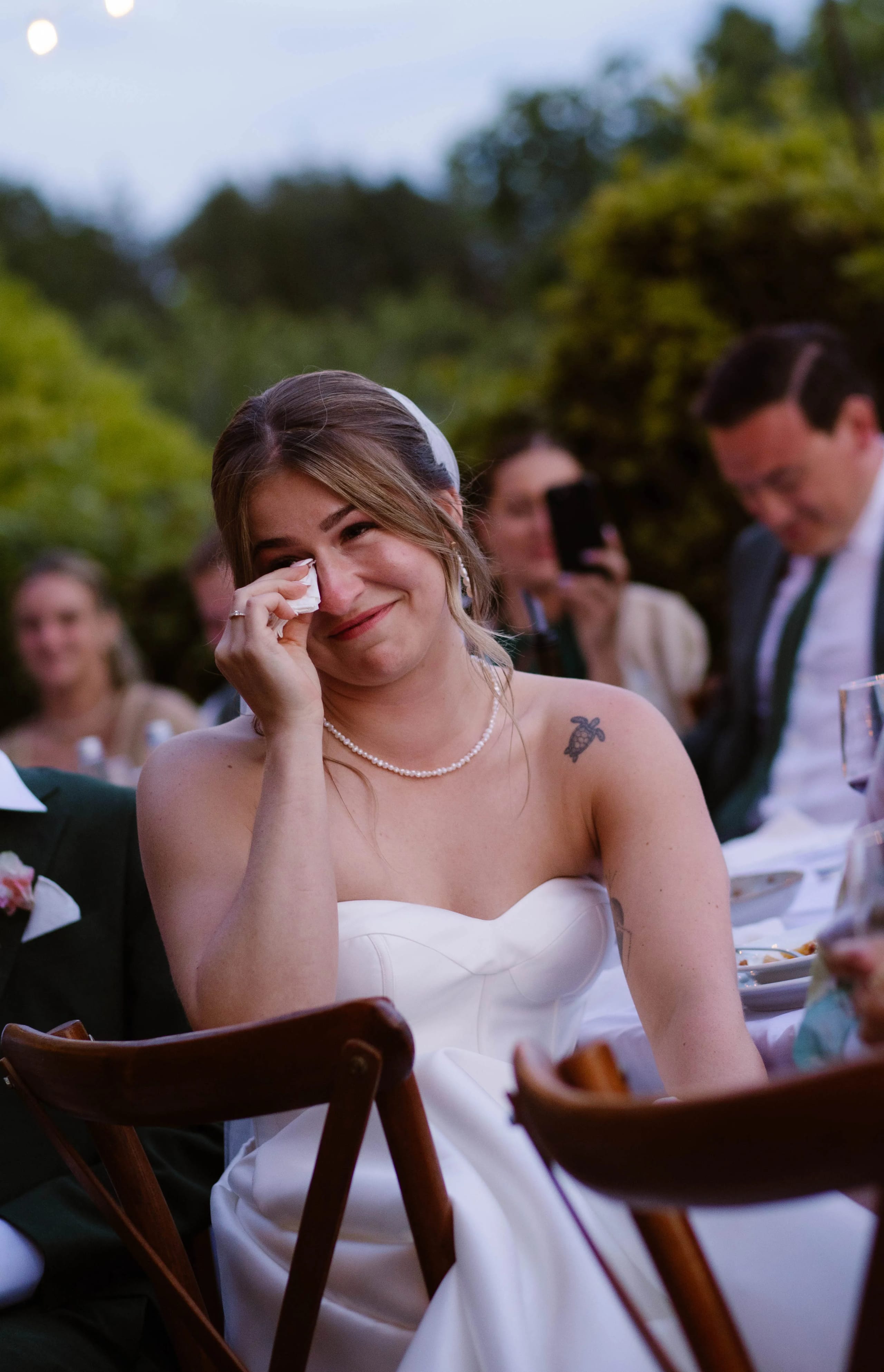 Een jonge vrouw in een witte trouwjurk veegt een traan weg terwijl ze emotioneel geniet van het huwelijksfeest, omringd door blije gasten in een mooie buitenomgeving.