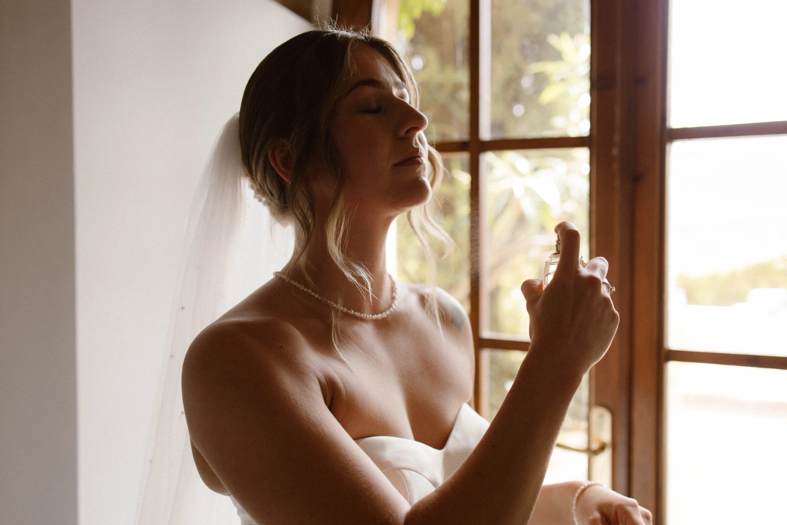 Een bruid in een strapless jurk sprayt parfum terwijl ze in stilte geniet van het moment bij een raam met uitzicht op een groene tuin.