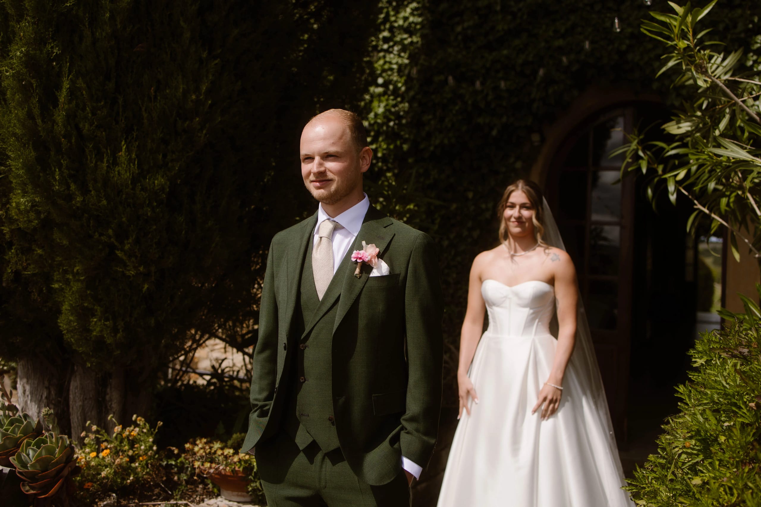 De bruidegom in een elegante groene pak staat met een zelfverzekerde houding voor een romantische achtergrond, terwijl de bruid in een stijlvolle witte trouwjurk zich op de achtergrond voorbereidt om hem te verrassen.