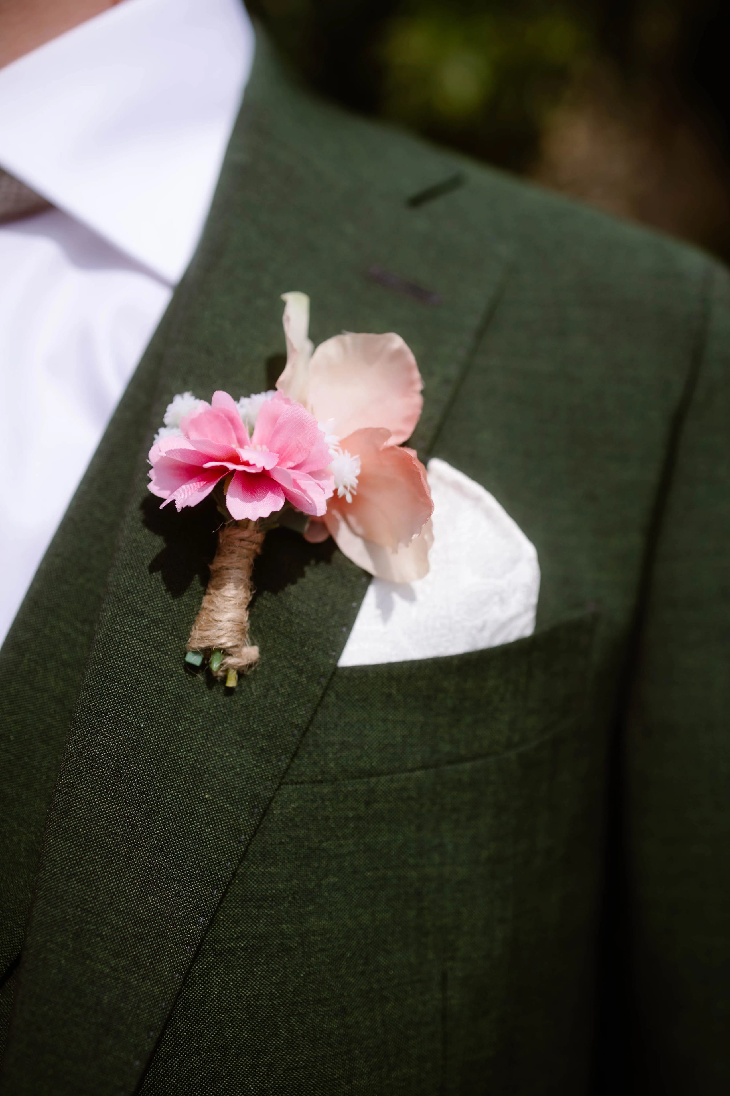 Een elegante bloemblaadje in zachte tinten van roze en wit, bevestigd aan de revers van een groen pak, met een witte zakdoek in de borstzak, perfect voor een formele gelegenheid.