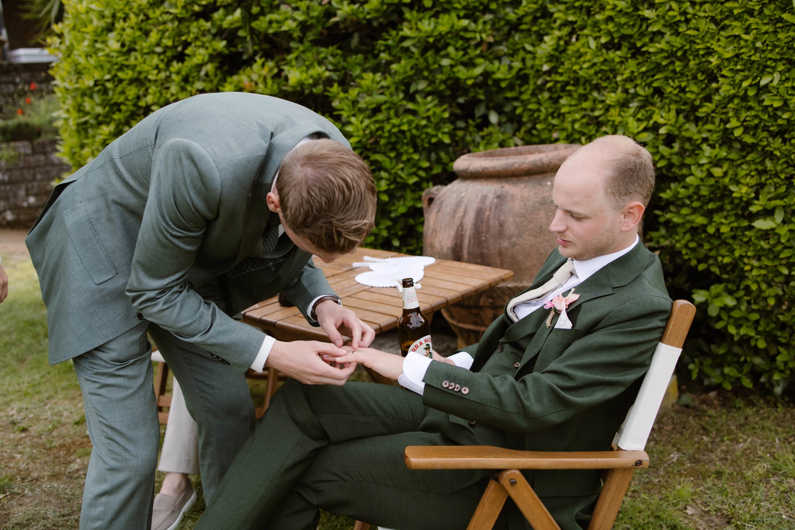 Een man in een groen pak helpt een andere man, ook in een groen pak, met het vastmaken van een strik terwijl ze op een houten stoel naast een tafel zitten, omringd door groen in een tuin.