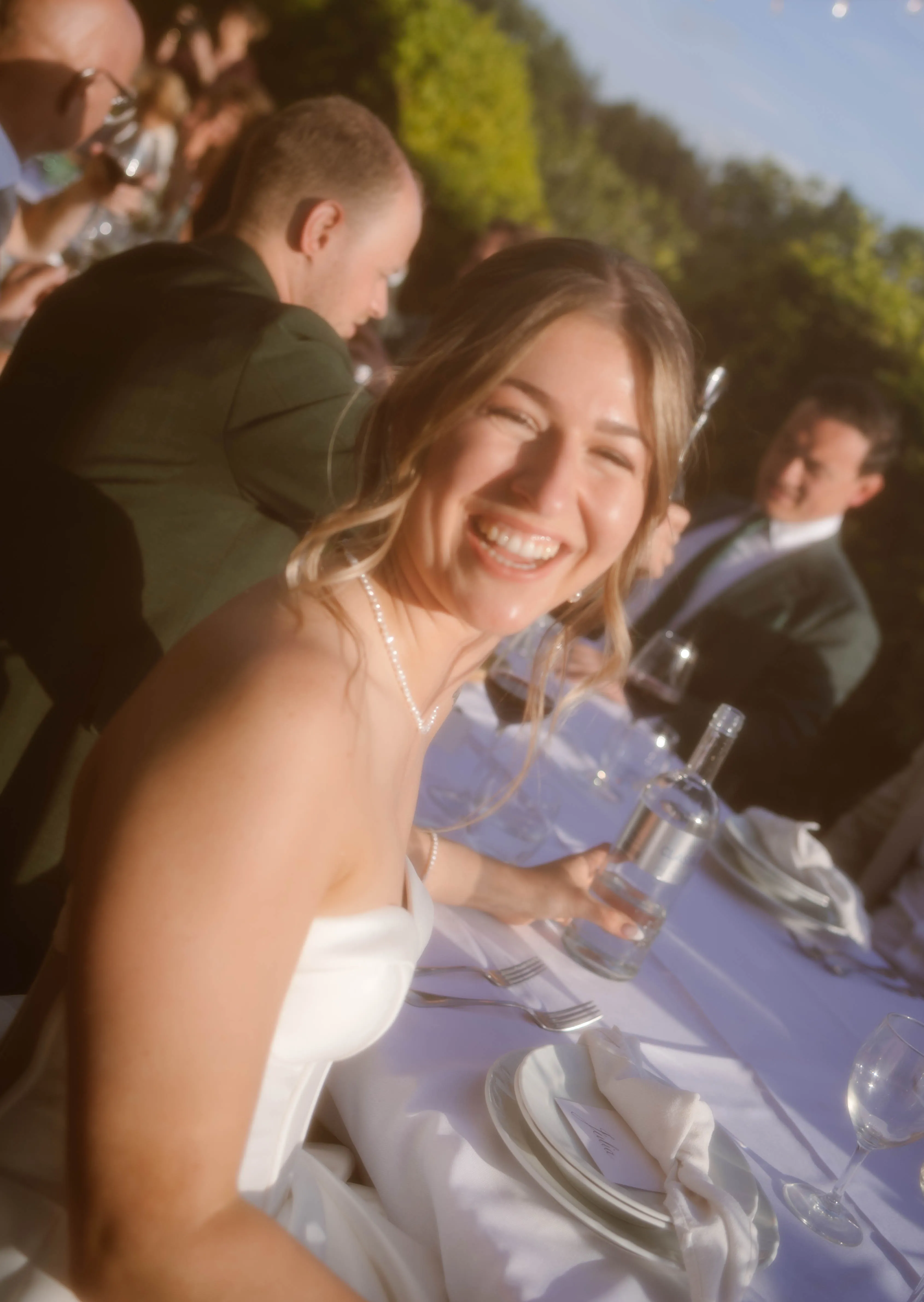 Een bruid in een strapless witte jurk glimlacht vrolijk terwijl ze aan een dinertafel zit, omringd door gasten in formele kleding, met een groene tuin op de achtergrond en sfeervolle verlichting.