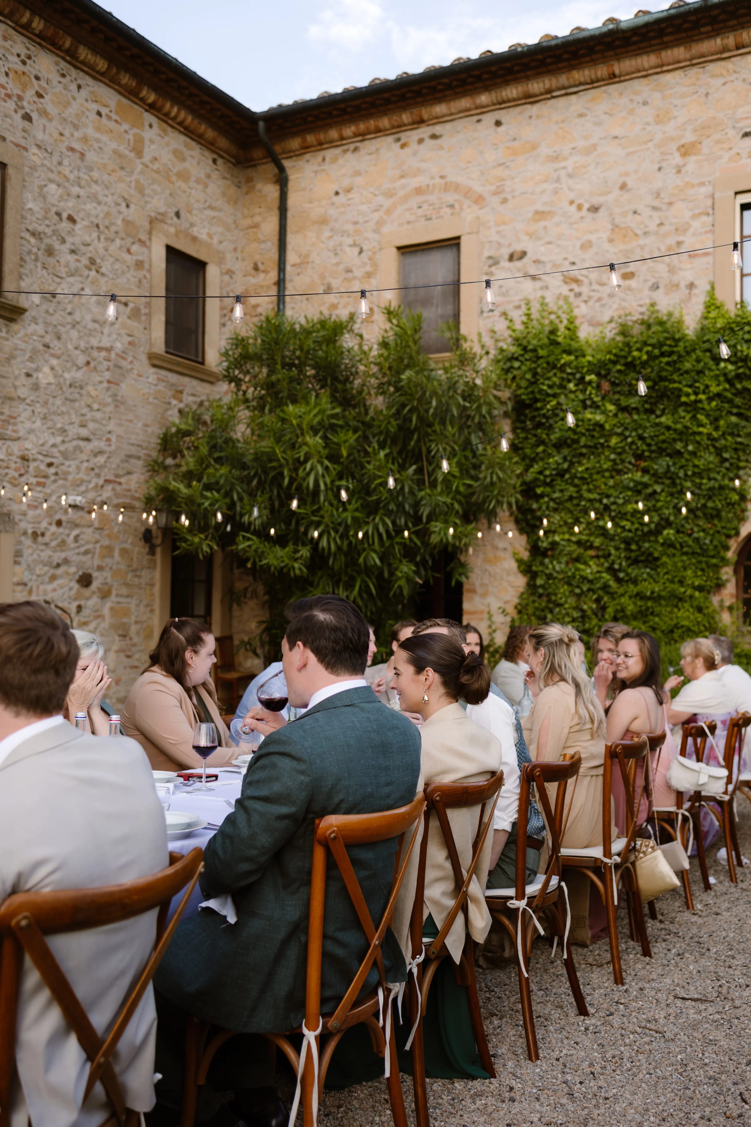 Een prachtig buiten diner met een lange tafel vol gasten, omringd door een charmante stenen muur, weelderig groen en sfeervolle verlichting in een rustige tuinsetting. De genodigden zijn in elegante kleding en genieten van het samenzijn in een feestelijke sfeer.