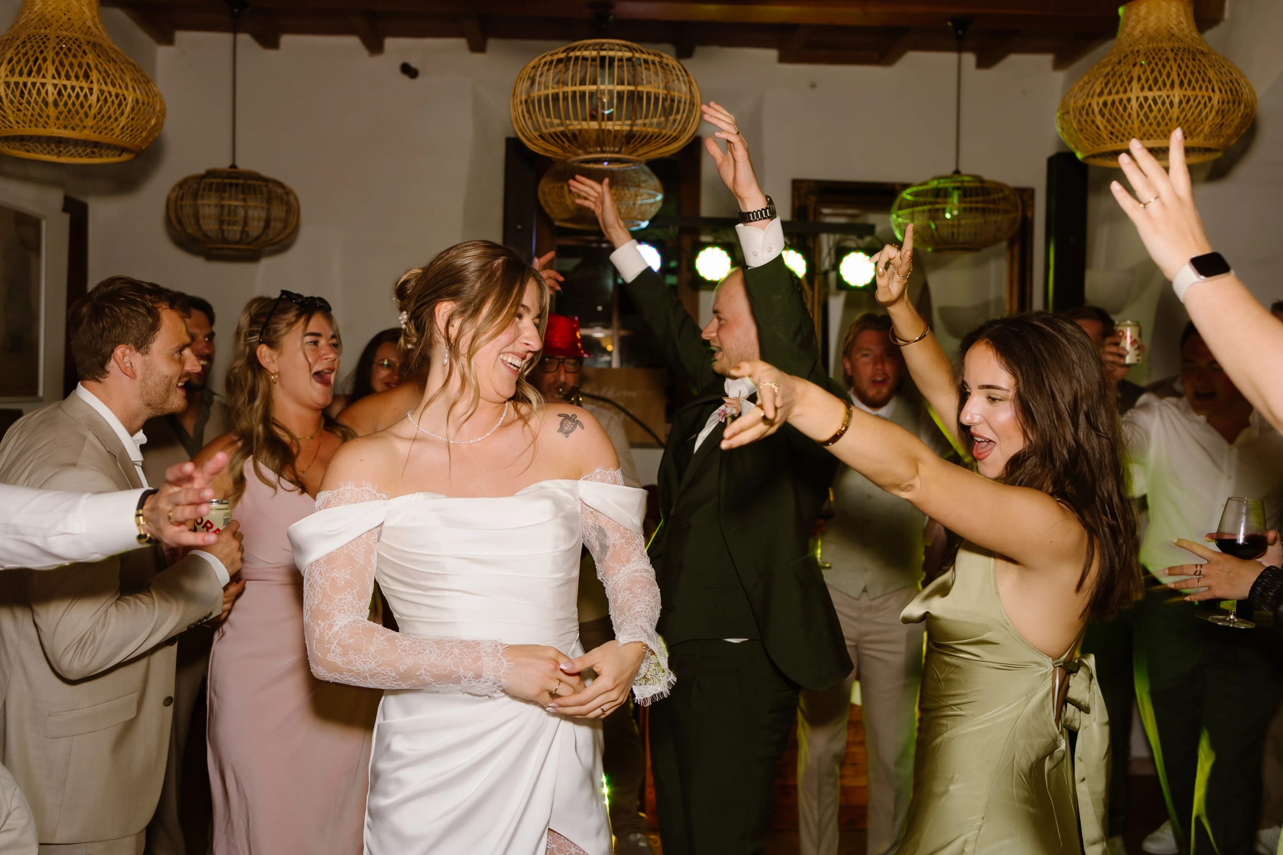 Een feestelijke dansvloer met verschillende mensen die vrolijk dansen en genieten van een viering, terwijl een bruid in een elegante witte jurk en een groep vrienden enthousiaste dansbewegingen maakt onder sfeervolle verlichting en decoratieve lampen.