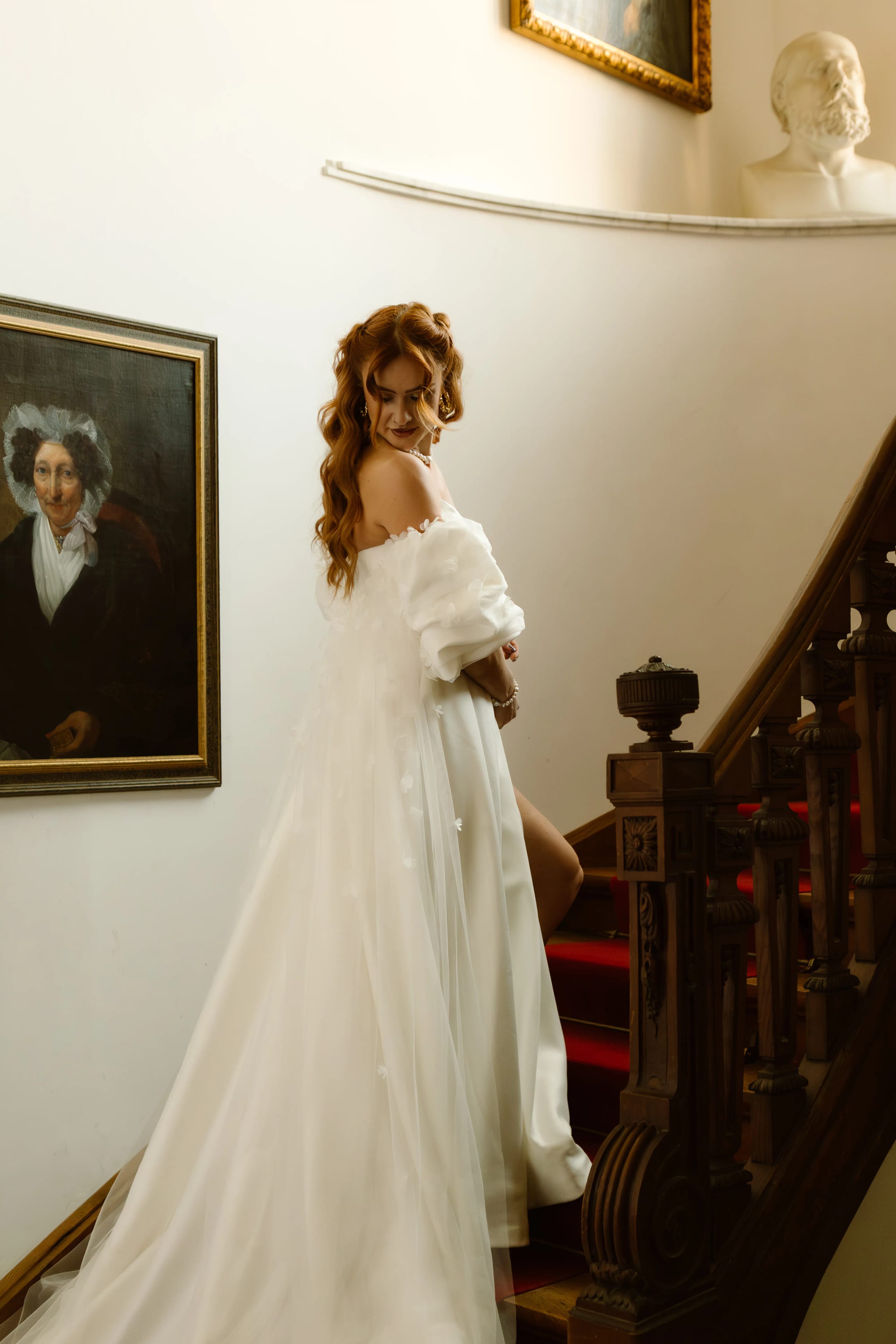 Een vrouw in een elegante witte trouwjurk poseert op een trap, met een schilderij van een historische vrouw en een bust van een man op de achtergrond, waardoor een sfeervolle, romantische setting ontstaat.