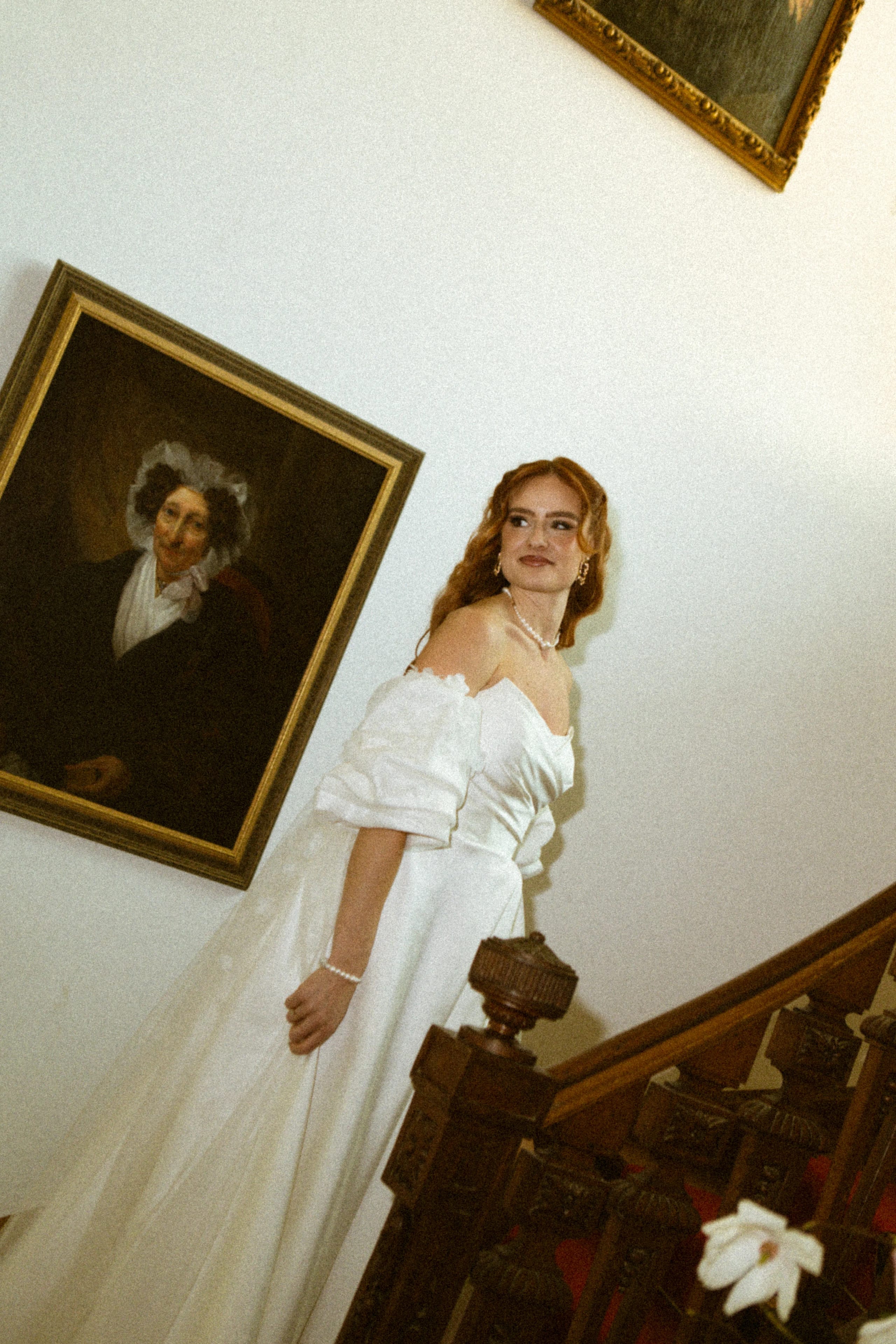 Een vrouw in een elegant wit trouwkleed staat op een trap, met een kunstwerk van een oudere man op de muur achter haar, wat een klassieke en romantische sfeer creëert. Haar lange, golvende haren en parelaccessoires accentueren haar verfijnde uitstraling.