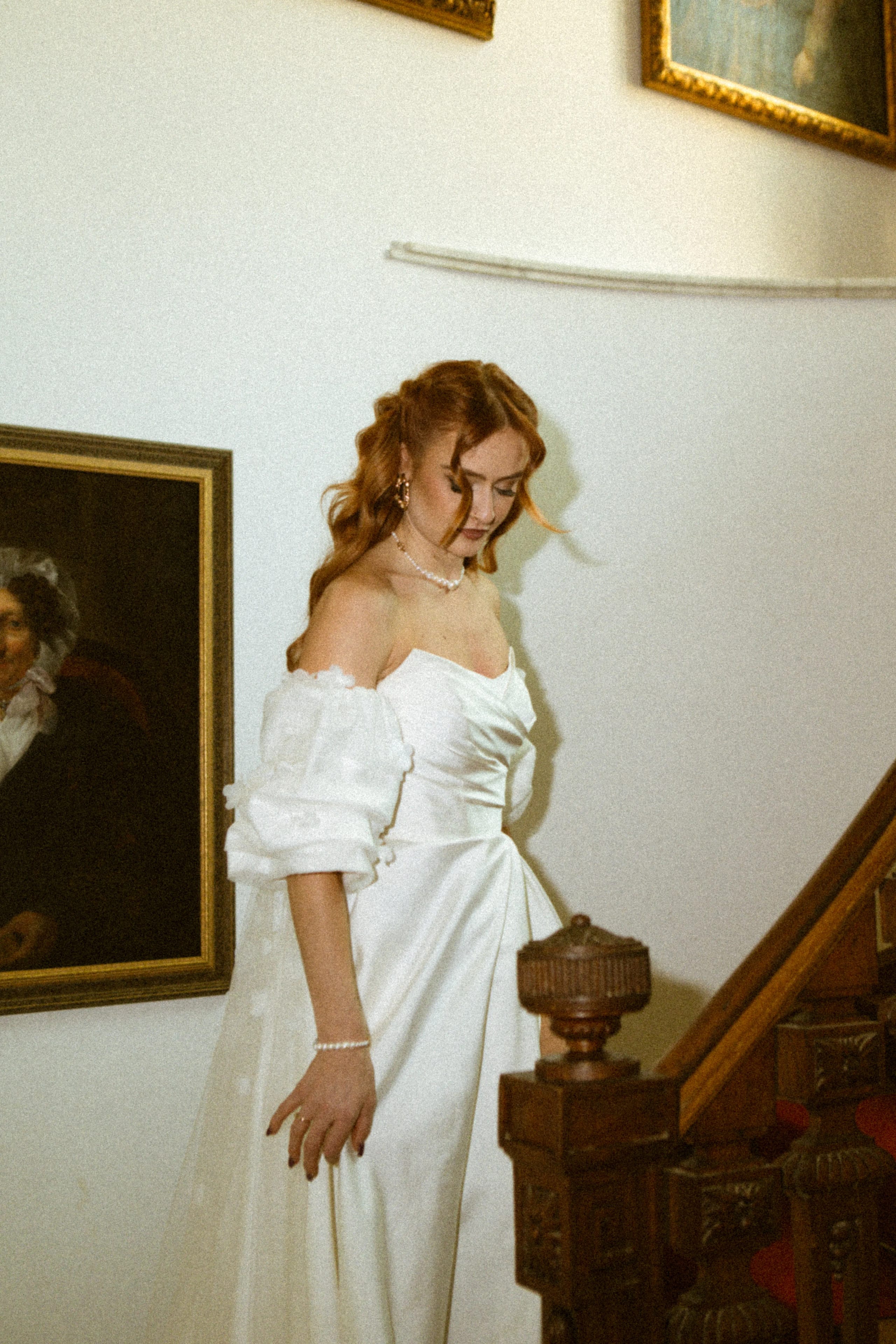 Een vrouw in een elegante witte jurk met pofmouwen en sieraden, terwijl ze voorzichtig een trap afloopt, omgeven door een klassieke interieur met historische schilderijen aan de muren.