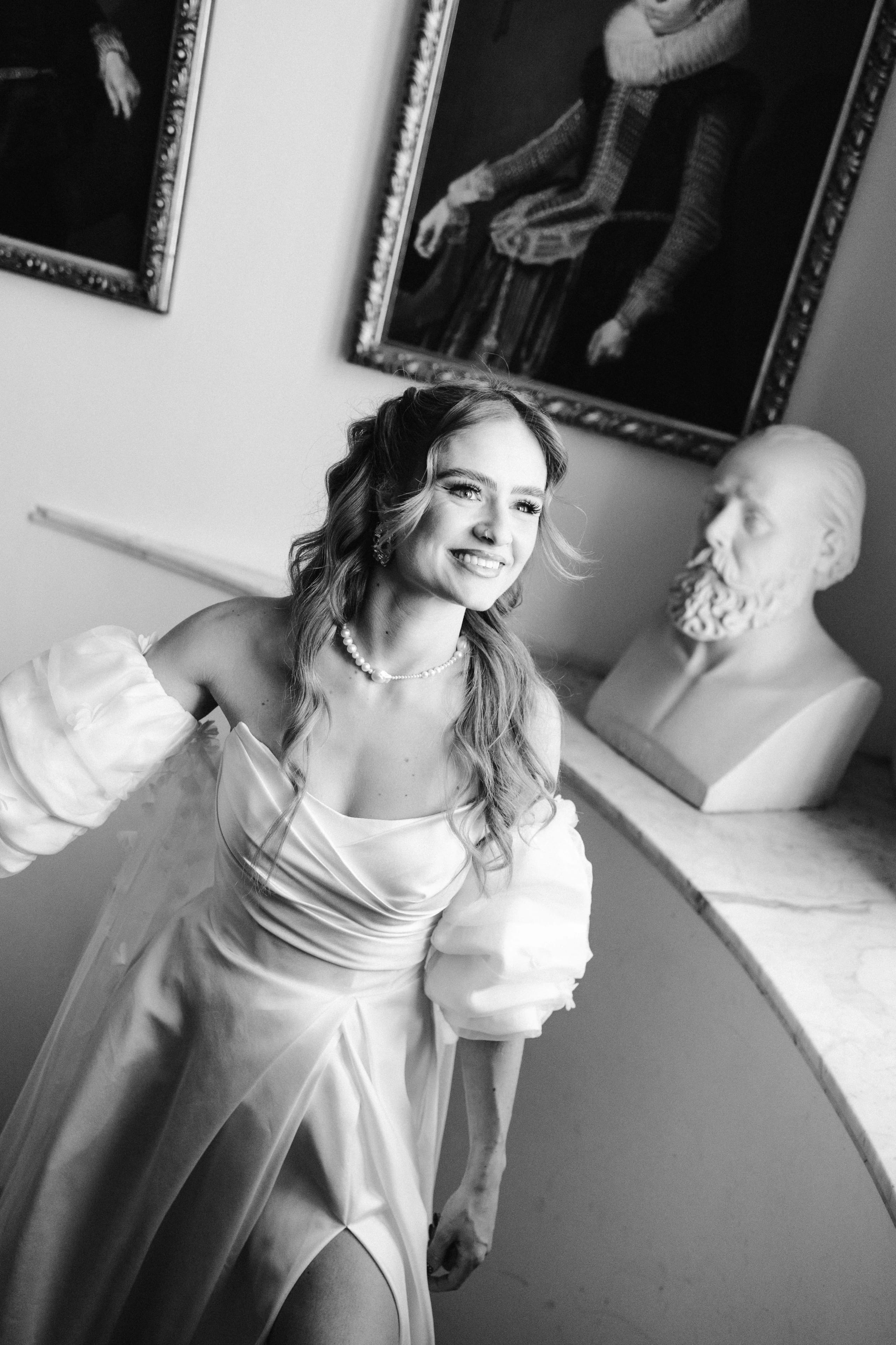 Een jonge vrouw met golvend haar draagt een elegante, lichte trouwjurk with schouderpuffen, terwijl ze glimlacht en poseert in een prachtig interieur met schilderijen en een klassiek standbeeld op de achtergrond.