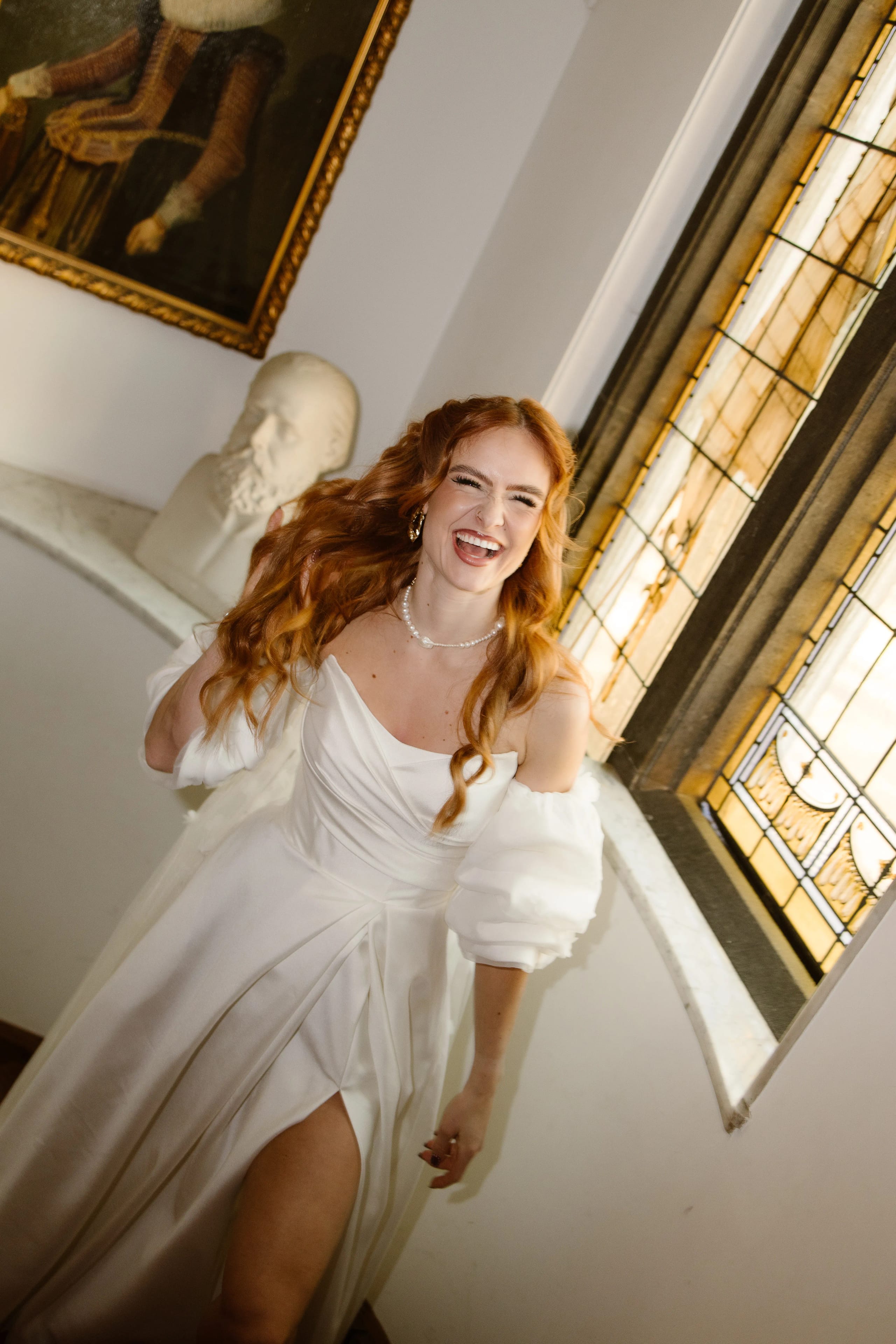 Een vrouw met lange, golvende kastanjebruine haren draagt een elegante witte jurk met een hoge split en schuimige mouwen, terwijl ze lachend poseert voor een raam met gebrandschilderd glas. Op de achtergrond is een bust van een klassieke kunstenaar te zien, wat een artistieke sfeer aan de ruimte toevoegt.