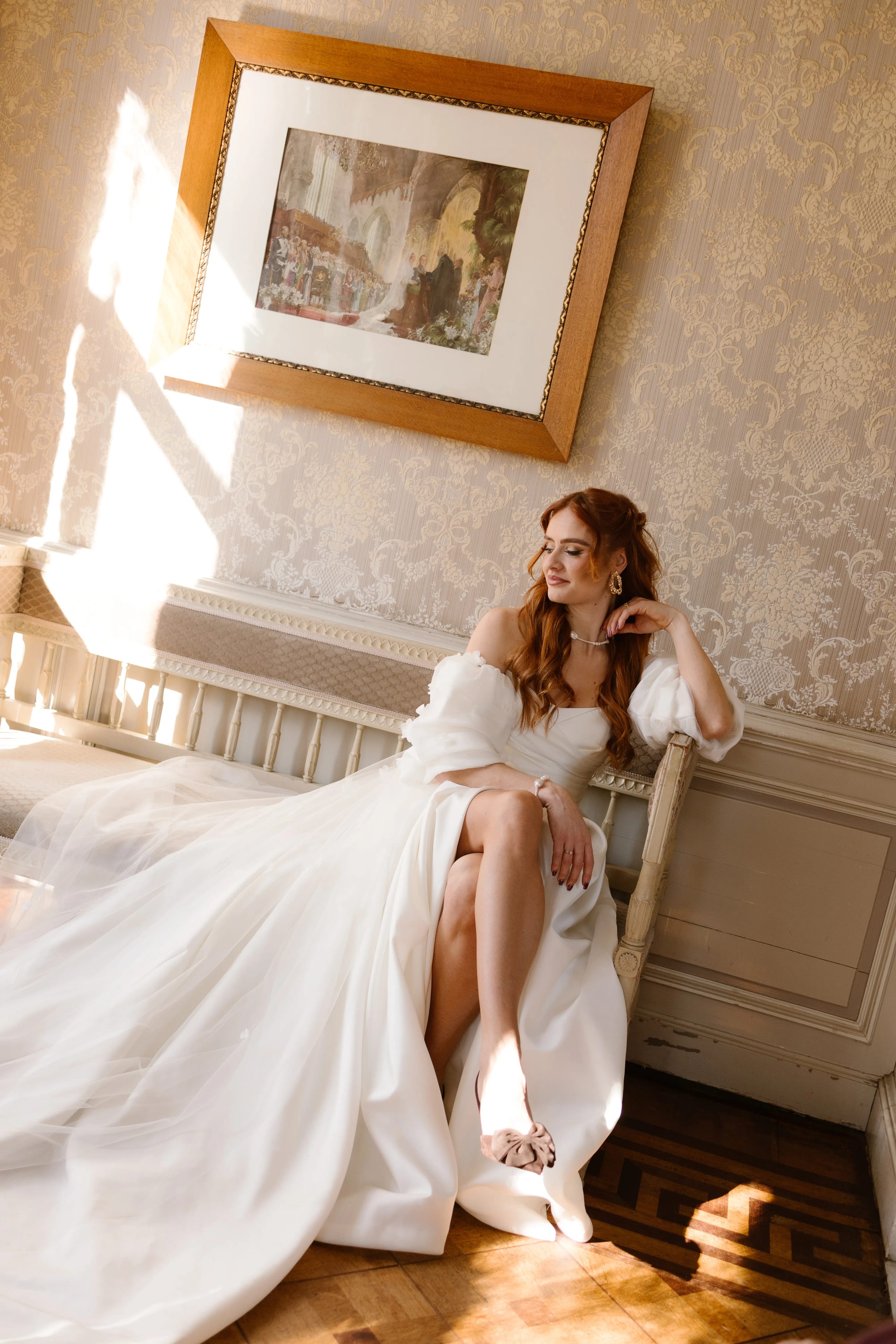Een stijlvolle bruid zit op een houten bank in een elegant interieur, gehuld in een prachtige witte trouwjurk met pofmouwen, verlicht door de zachte zonneschijn die door het raam valt. Achter haar hangt een kunstwerk, wat een luxe ambiance toevoegt aan de scène.