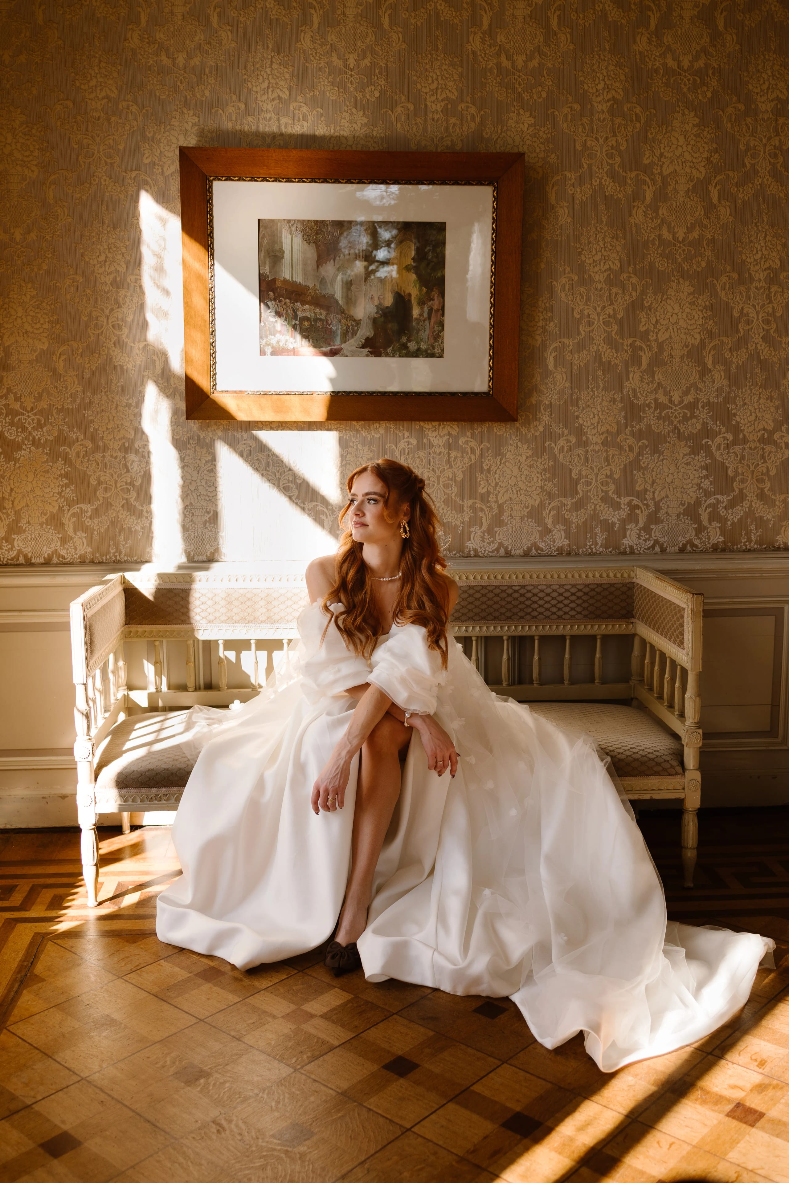 Een elegante bruid in een romantische trouwjurk zit op een houten bank in een sfeervolle ruimte, omringd door warm licht dat door het raam valt, met een kunstwerk aan de muur op de achtergrond.