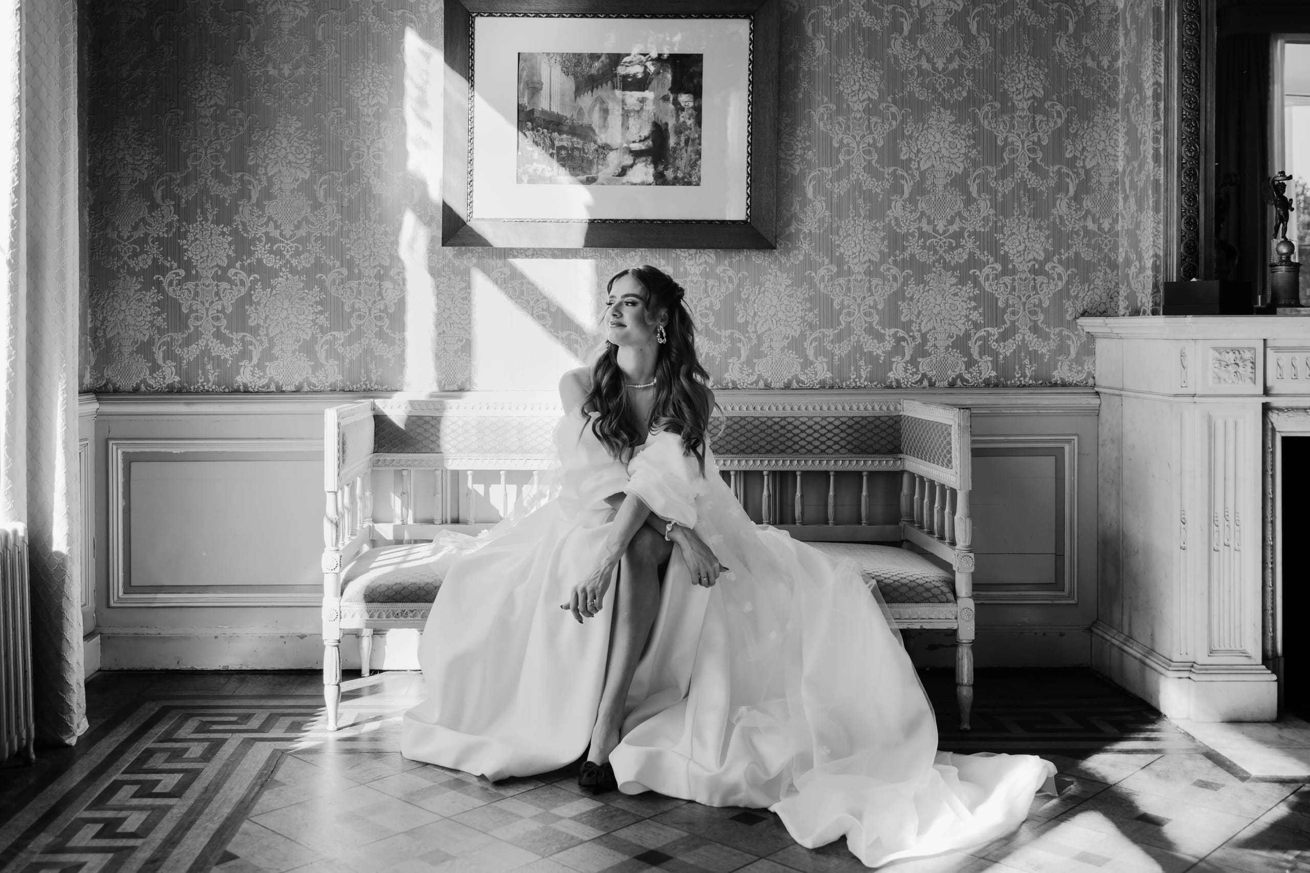 Een bruid in een elegant wit trouwkleed zit op een houten bank in een klassiek ingerichte kamer, omringd door sfeervol licht dat door het raam valt, terwijl ze een dromerige blik heeft. De stijlvolle achtergrond met tapijten en een kunstwerk voegt charme toe aan het tafereel.
