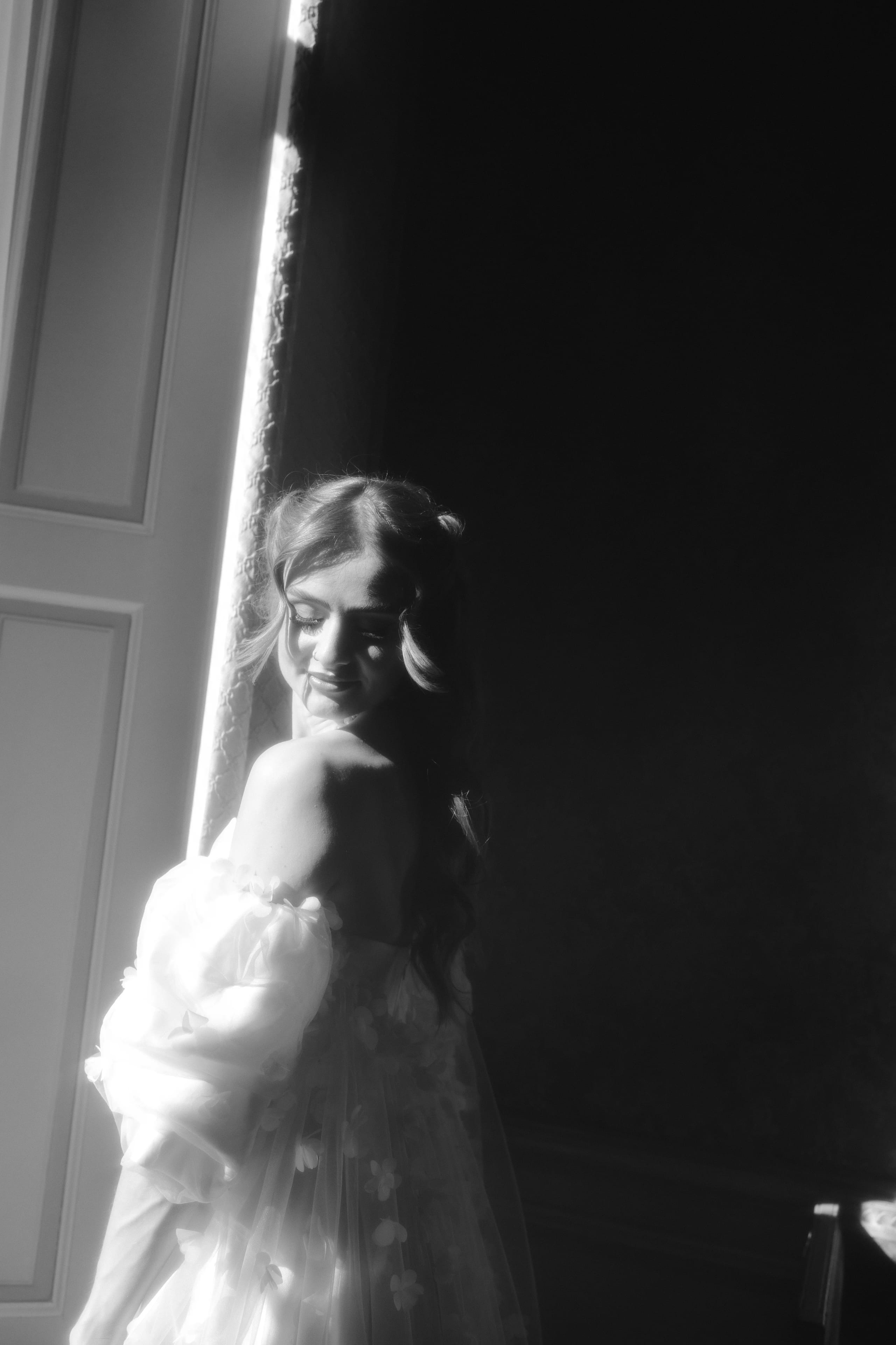 Een vrouw in een romantische, doorzichtige jurk met bloemendetails kijkt over haar schouder, badend in zacht natuurlijk licht dat door een open deur valt, waardoor een dromerige sfeer ontstaat.