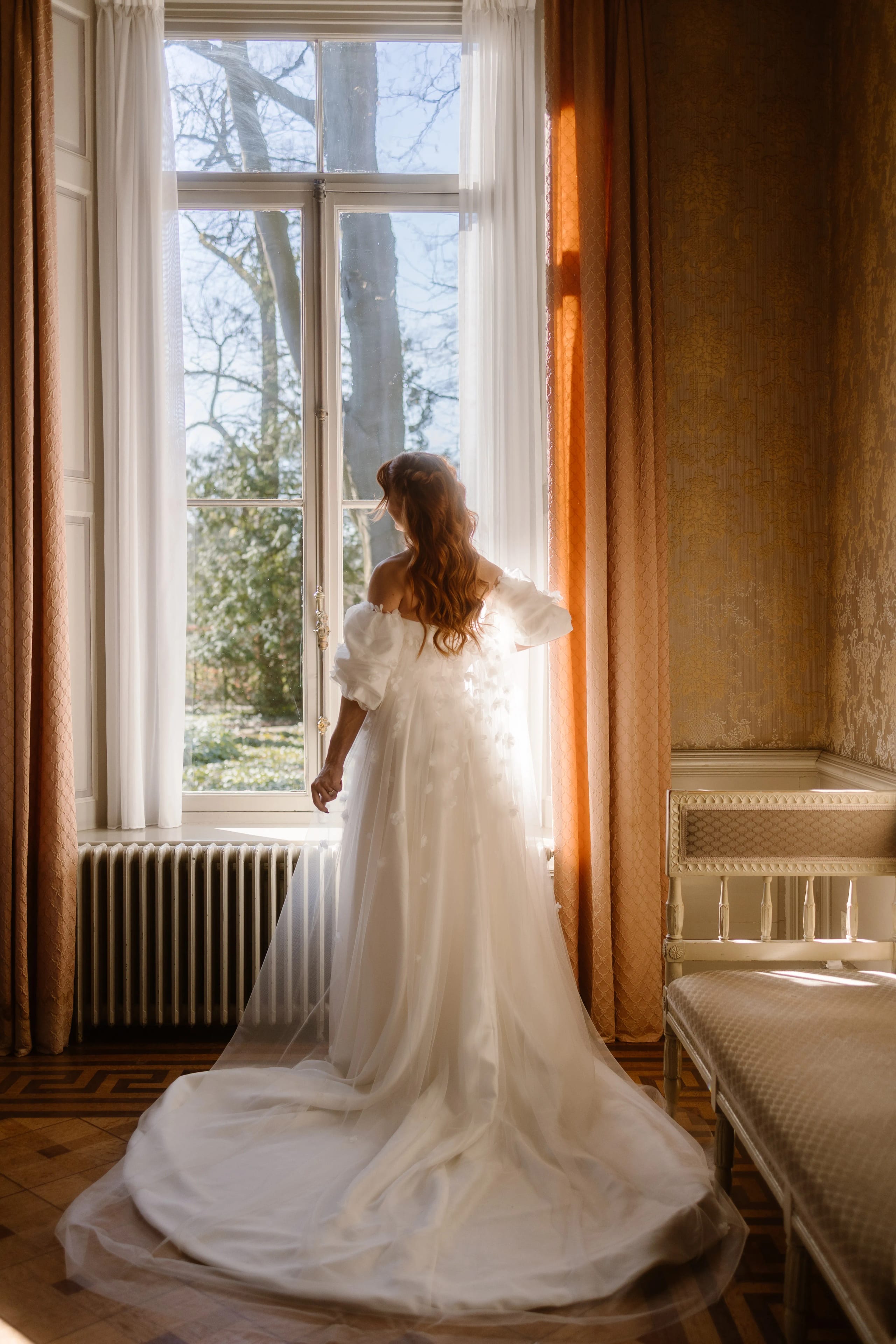 Een bruid in een elegante witte trouwjurk met een lange sleep staat bij een grote raam met gordijnen, terwijl ze naar buiten kijkt op een zonnige dag, omringd door de sfeervolle inrichting van een historisch interieur.