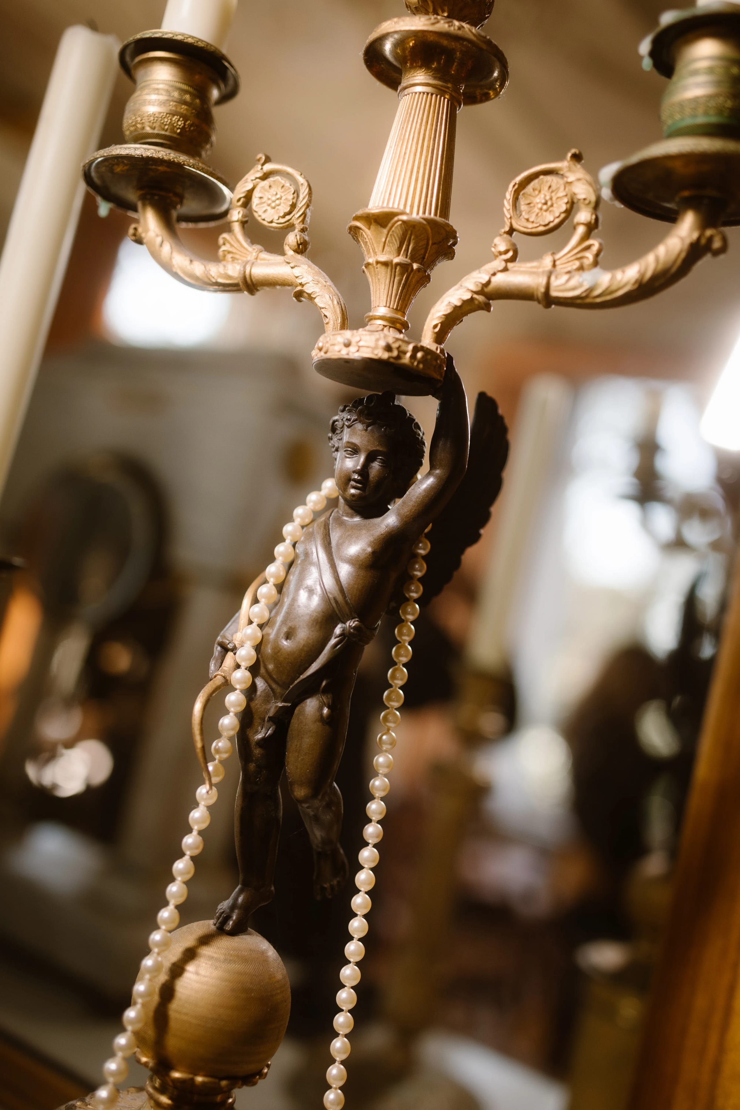 Een gedetailleerde bronzen kandelaar met een schattige engel die aan de onderkant hangt, omringd door parelkettingen, en een warme achtergrond met vintage decor.