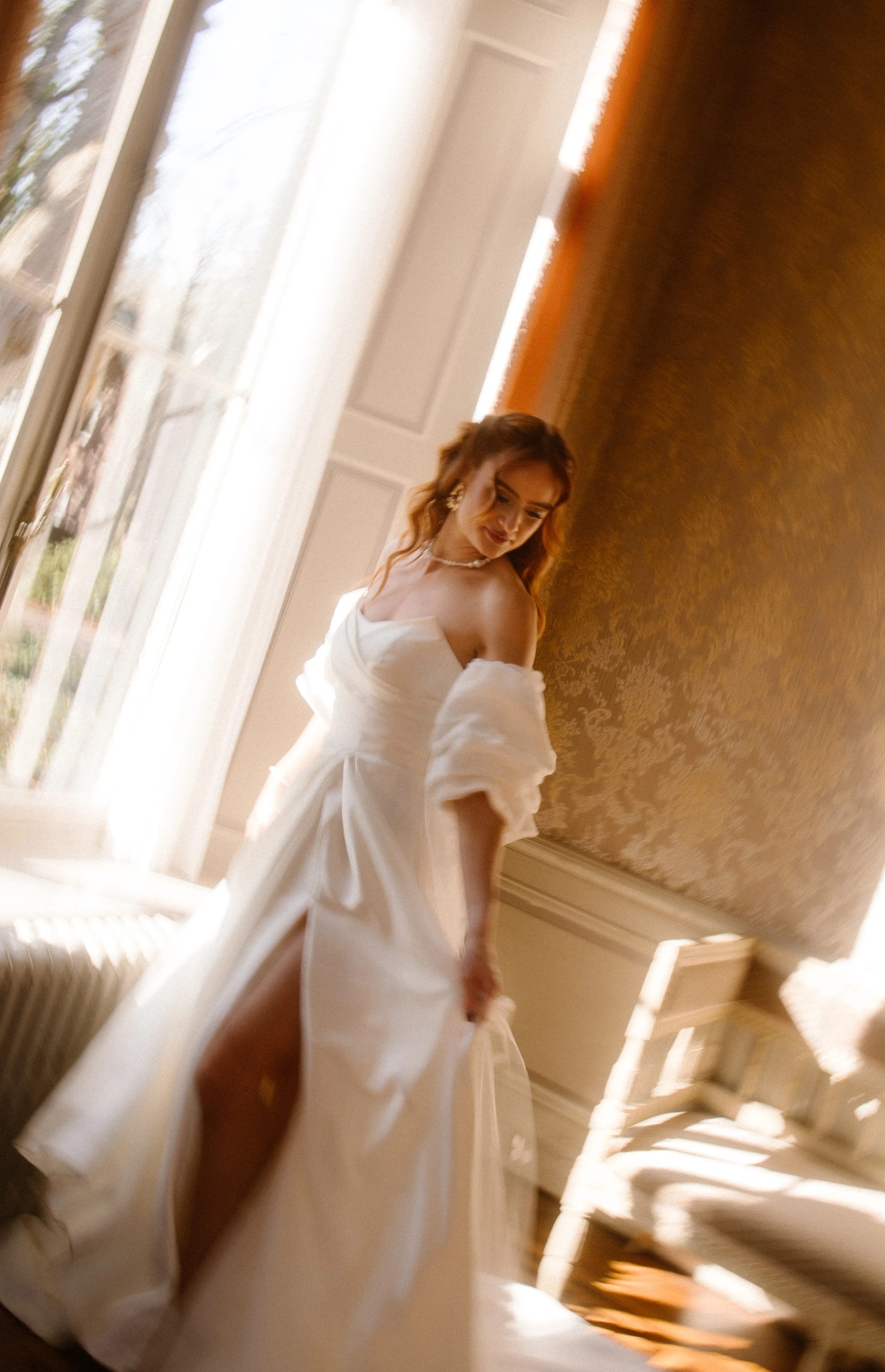 Een bruid in een elegante witte jurk met mooie schouders, die zich speels beweegt in een zonnige kamer met grote ramen, waardoor een romantische sfeer ontstaat. De zachte verlichting benadrukt de details van de jurk en de sierlijke uitstraling van de bruid.
