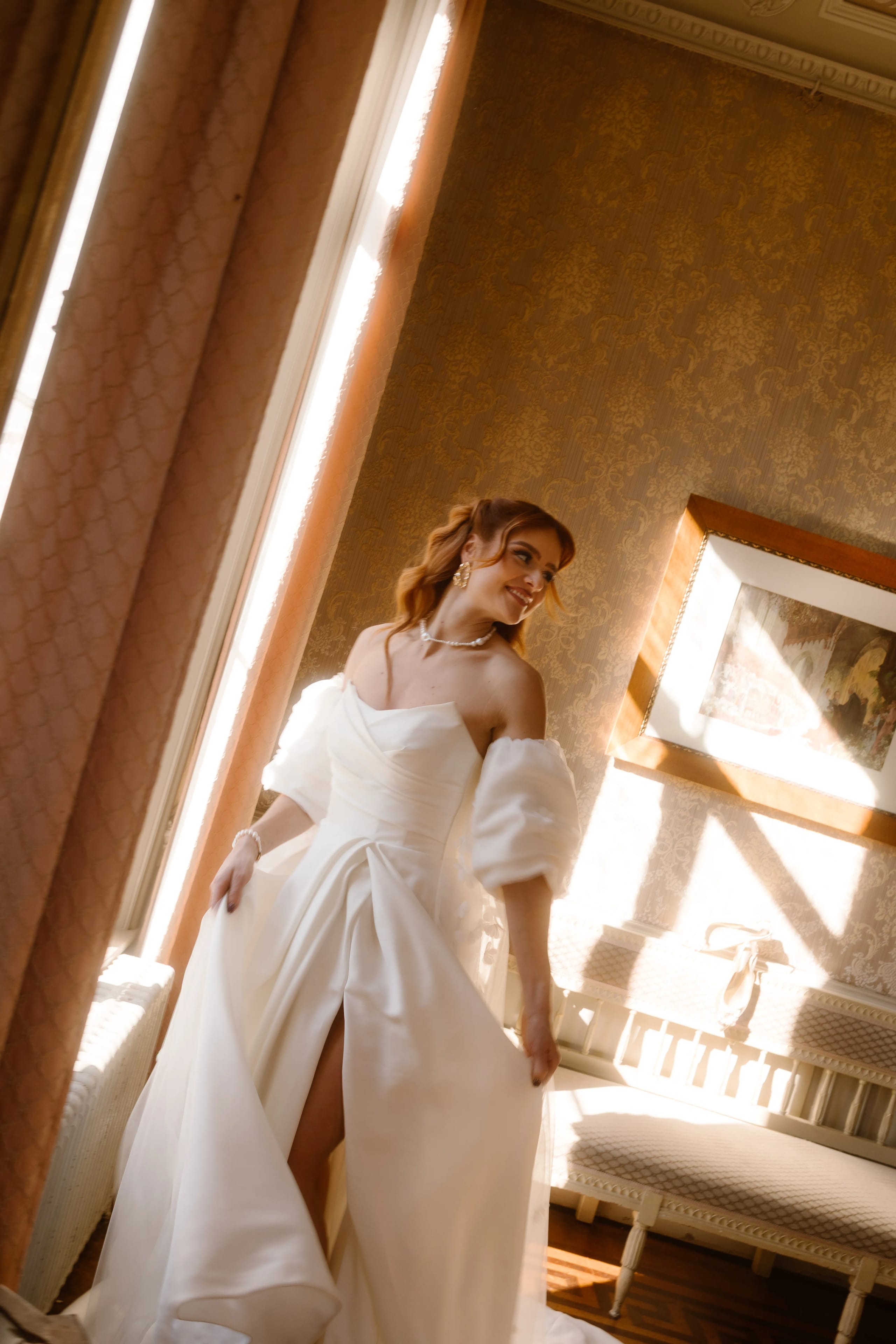 Een bruid in een elegante witte trouwjurk met pofmouwen poseert stralend in een kamer met warm licht en vintage inrichting, omringd door een warme, sfeervolle ambiance.