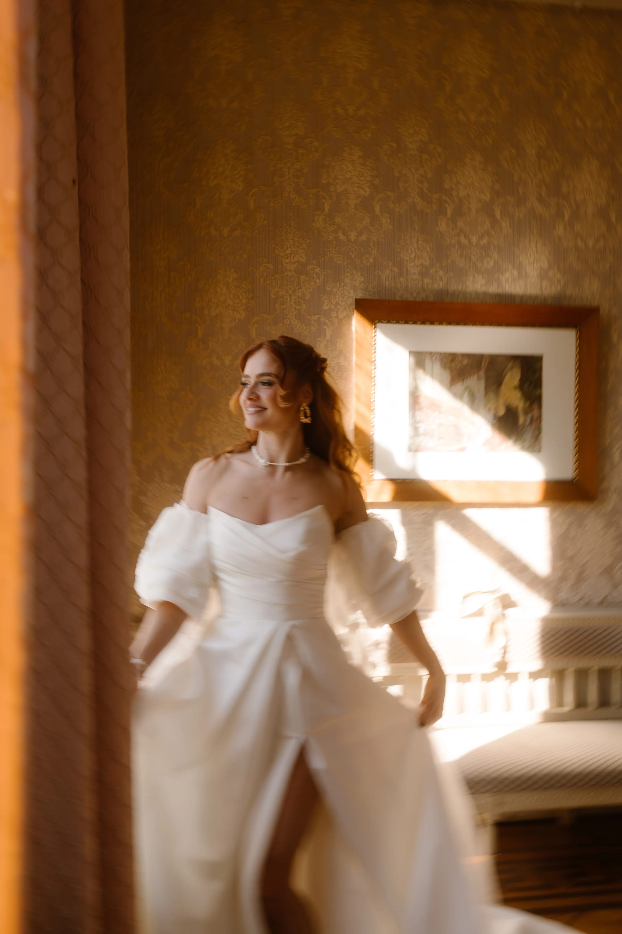 Een bruid in een elegante, witte trouwjurk met pofmouwen, terwijl ze met een glimlach door een zonverlichtere kamer beweegt, omringd door een warme, vintage sfeer.