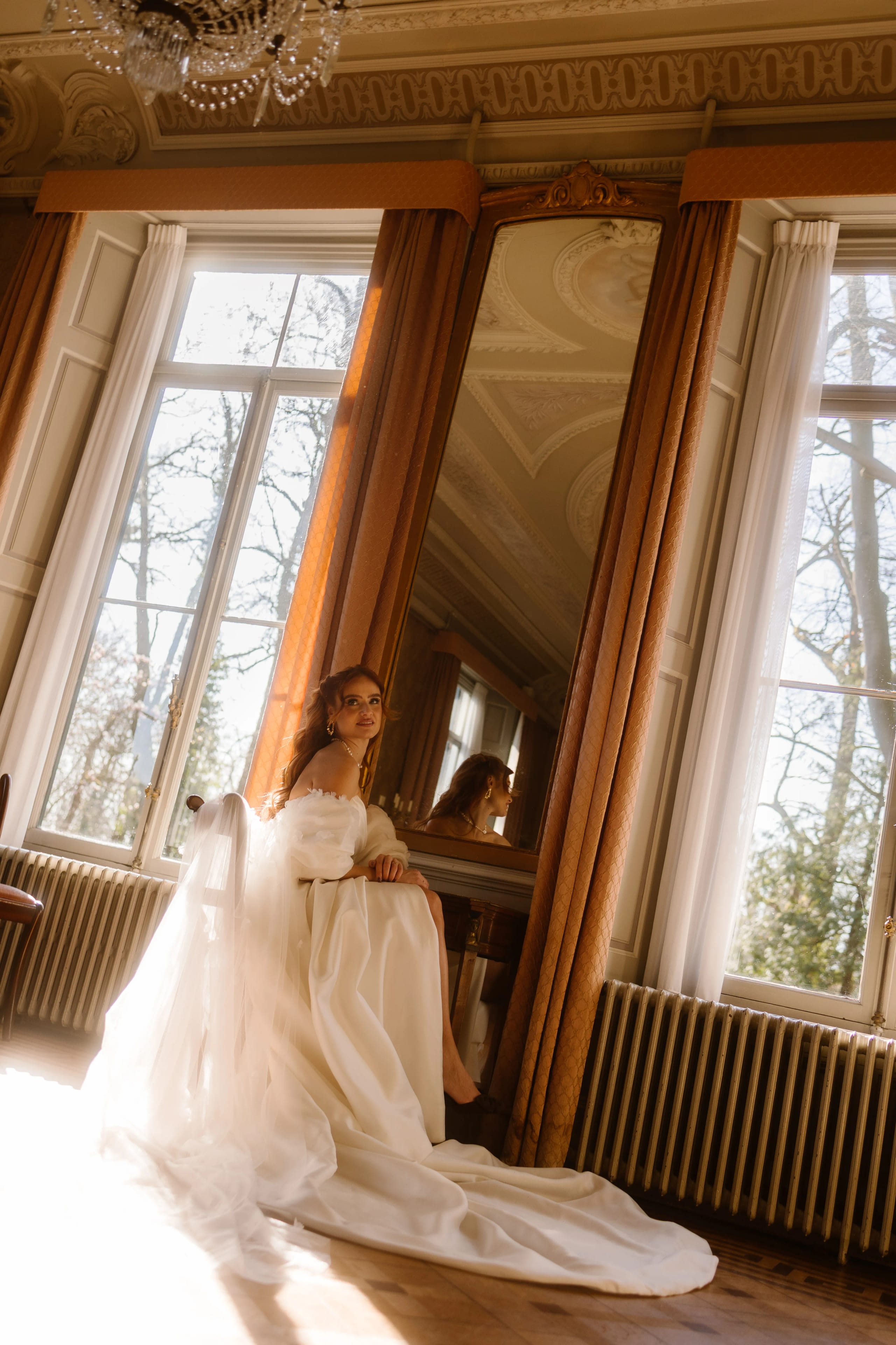 Een bruid in een elegante trouwjurk zit op de vloer van een licht verlichte kamer met grote ramen en lange, mooie gordijnen. Haar hoofd is gekanteld terwijl ze naar haar spiegelbeeld kijkt, omgegeven door een statige ambiance en details van klassieke decoratie.