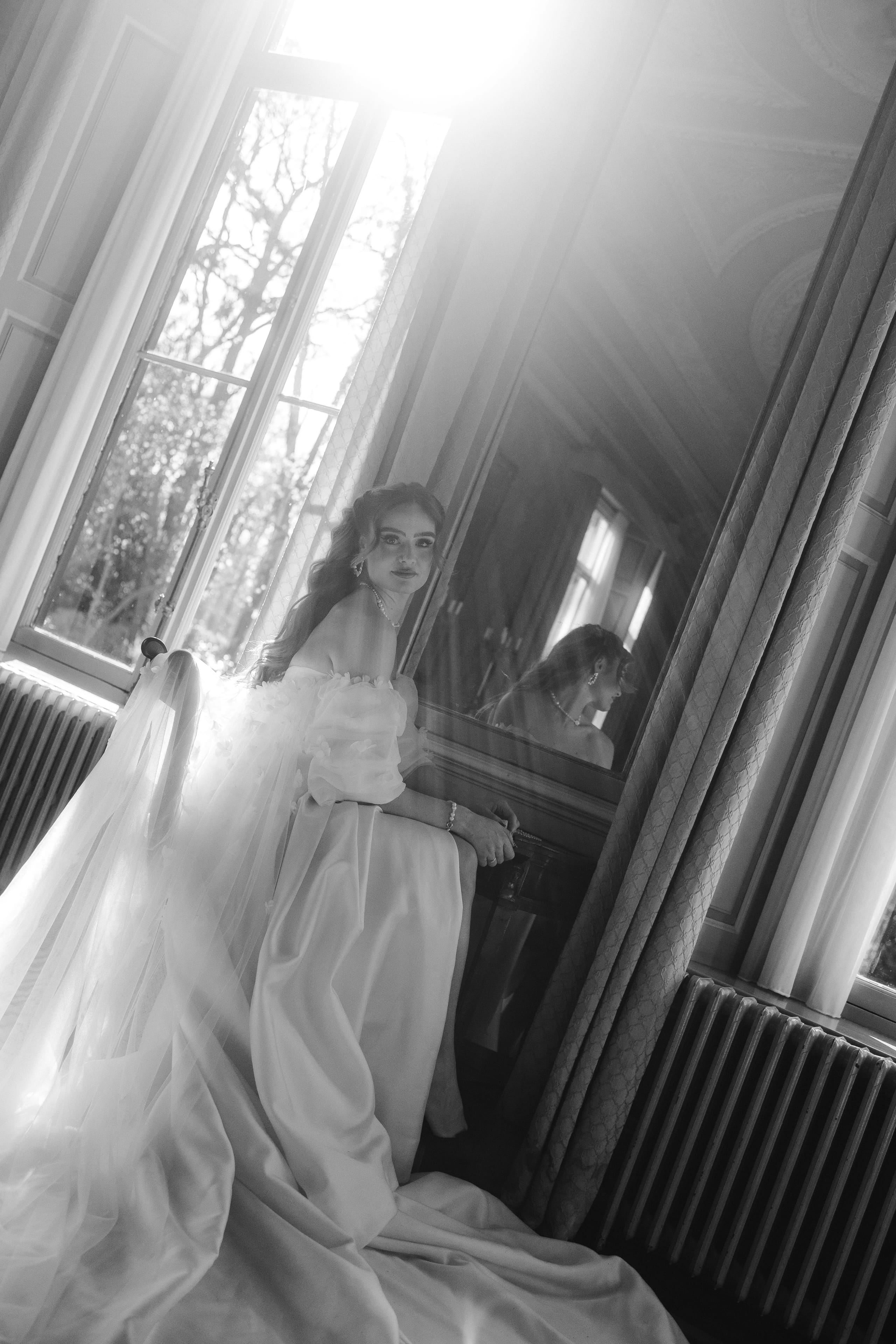Een vrouw in een elegante, witte jurk poseert in een sfeervolle kamer met grote ramen, terwijl het zonlicht door de gordijnen naar binnen valt en een reflectie in de spiegel zichtbaar is. De afbeelding straalt een romantische en dromerige sfeer uit, met een vintage uitstraling door het zwart-wit effect.