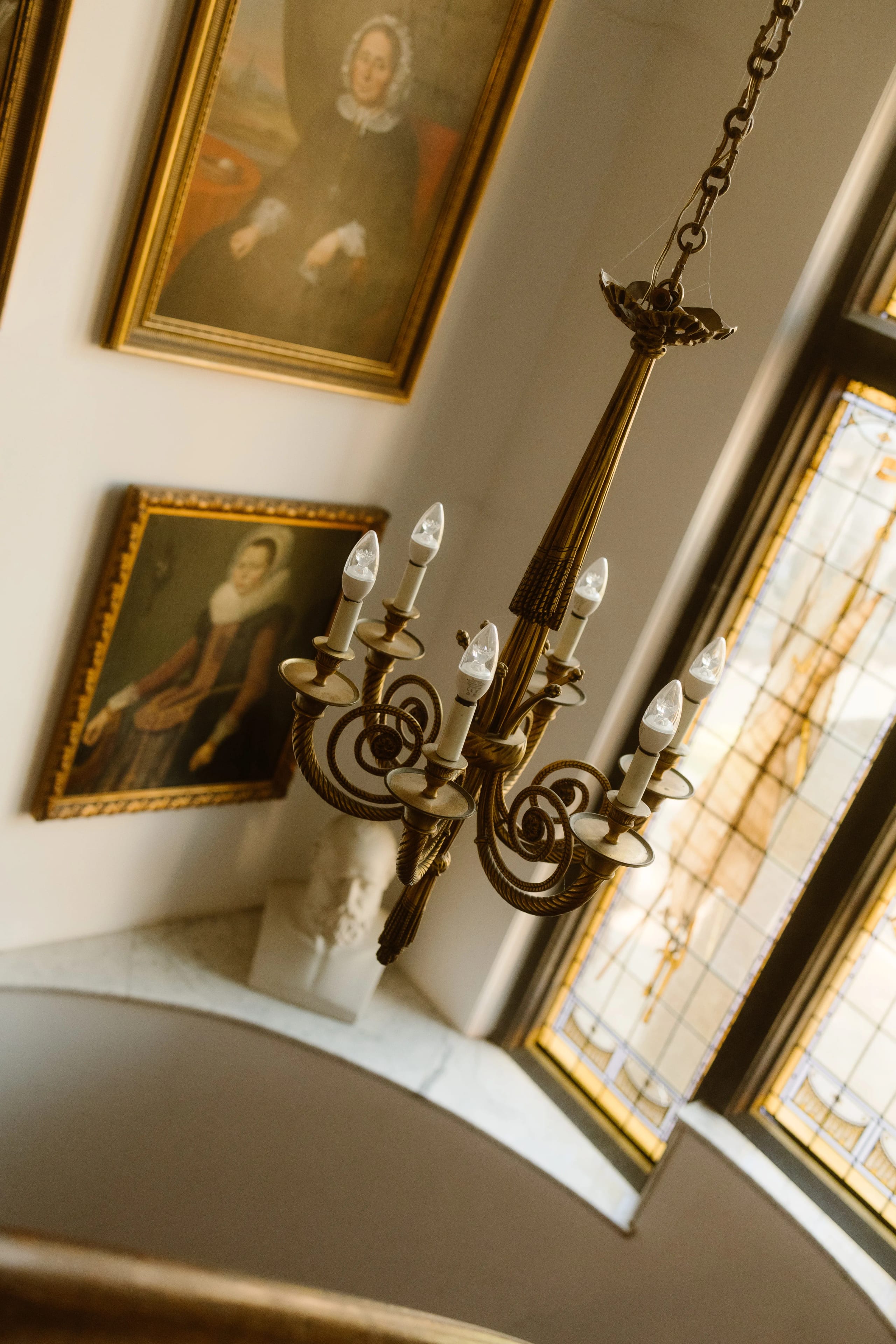 Luxe kroonluchter met elegante kaarslampen hangt boven een stijlvolle trap, omringd door klassieke schilderijen en een stenen beeld, verlicht door natuurlijk licht dat door de glas-in-loodramen valt.