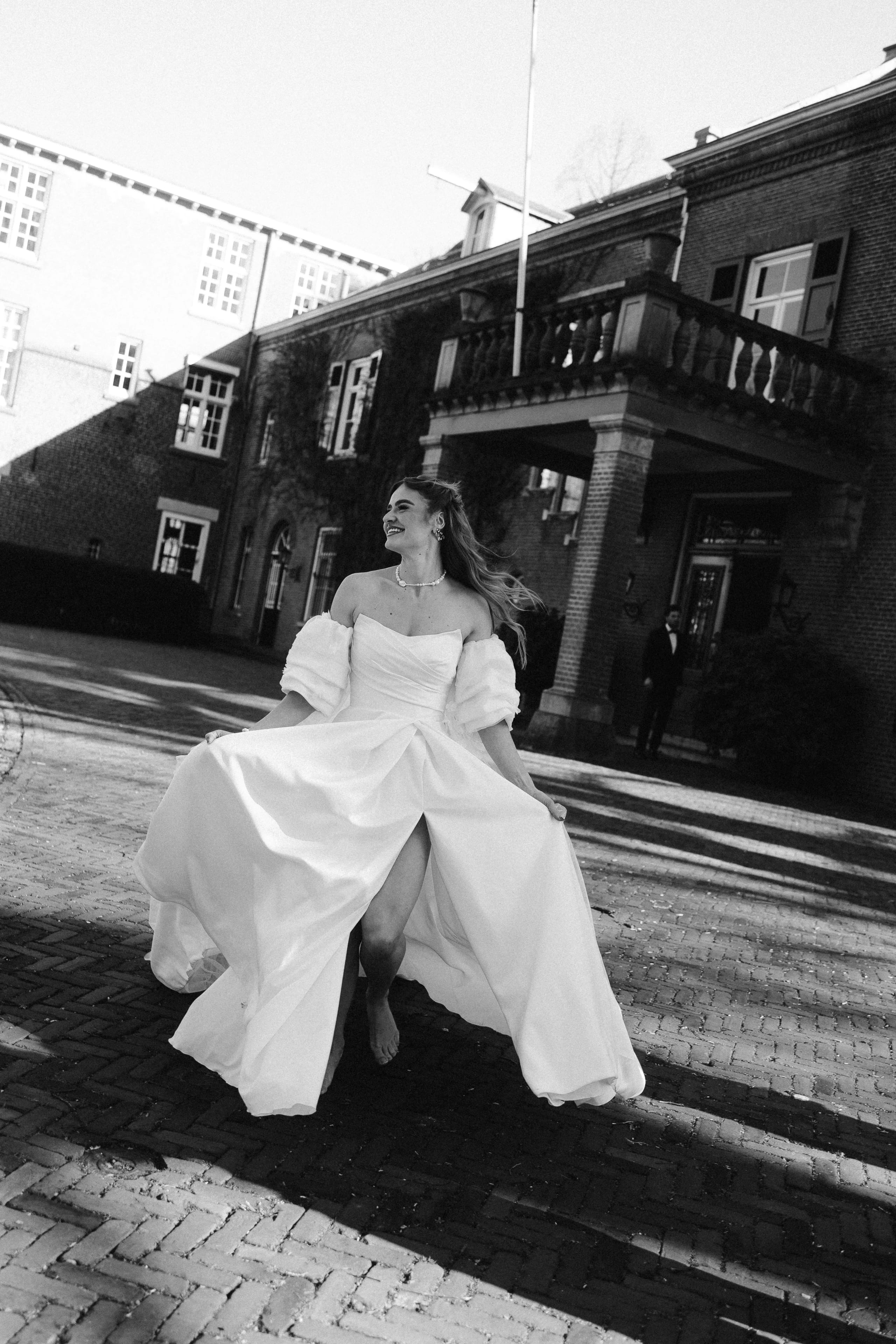 Een bruid danst met een stralende lach in een elegante witte jurk met pofmouwen, terwijl ze zich op een zonnige dag op een charmante kasseistraat bij een statig gebouw bevindt.