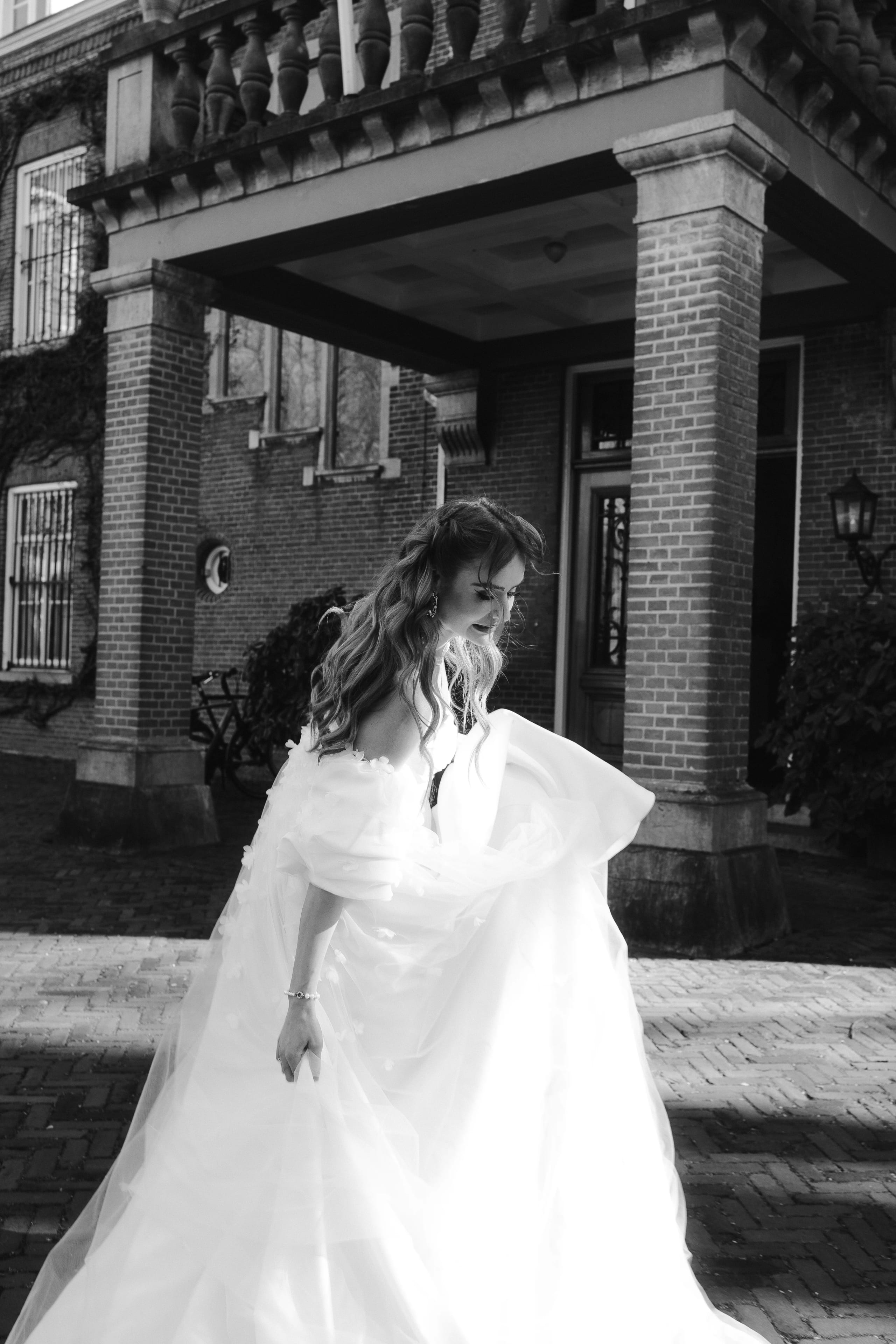 Een bruid in een weelderige witte jurk wandelt vrolijk in de schaduw van een statig pand, terwijl haar jurk sierlijk om haar heen waait.