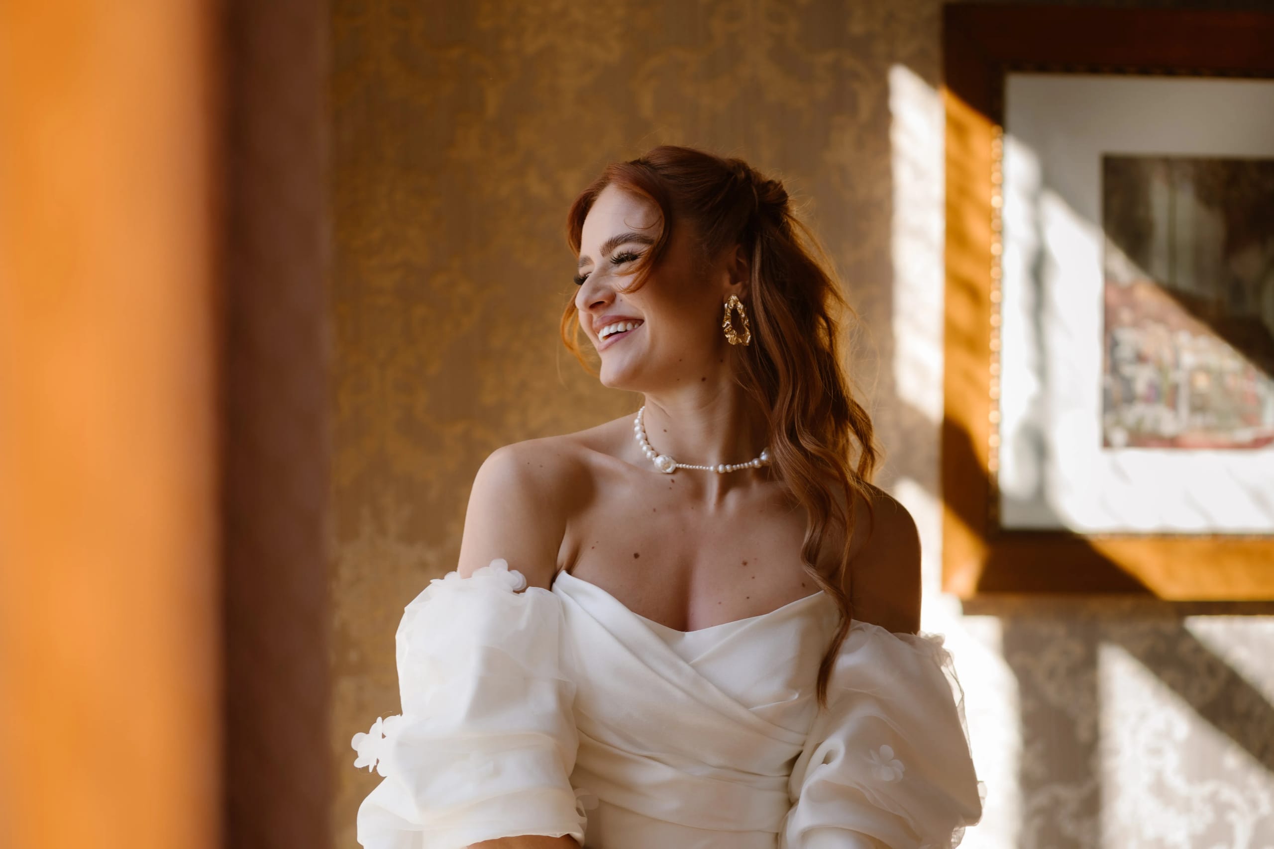 Een stralende vrouw in een elegante witte jurk met voluminieuze mouwen, zittend bij een raam met gouden verlichting, terwijl ze glimlacht en haar kapsel met zachte golven bewondert. Haar sieraden, waaronder een parelketting en grote oorbellen, voegen een luxe tintje toe aan haar uitstraling.