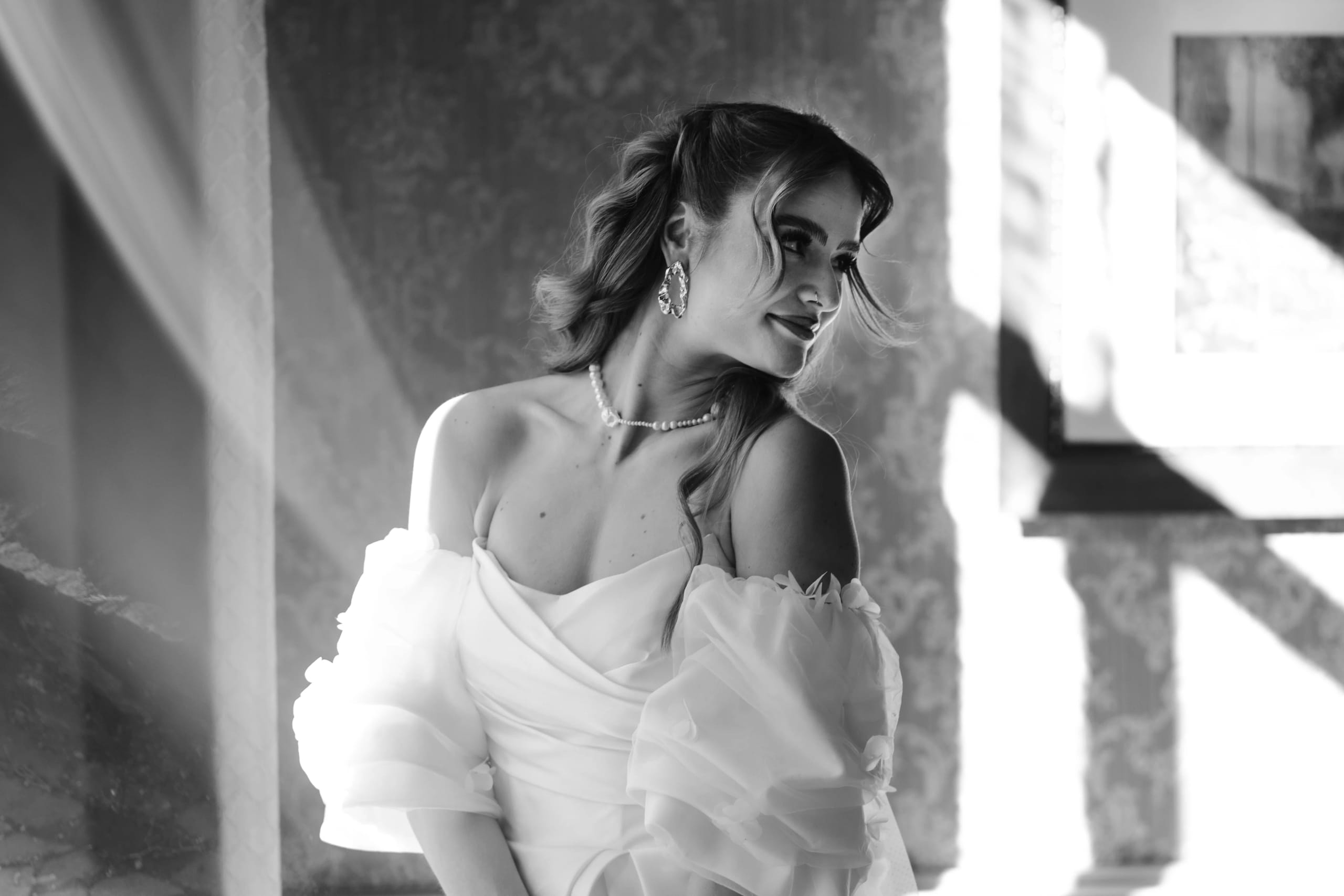 Een jonge vrouw in een elegante witte trouwjurk, met pofmouwen, kijkt charmant over haar schouder in een sfeervolle omgeving, verlicht door zachte stralen van natuurlijk licht. Haar kapsel en accessoires, zoals grote oorbellen en een parelketting, voegen een verfijnde uitstraling toe aan de romantische sfeer.