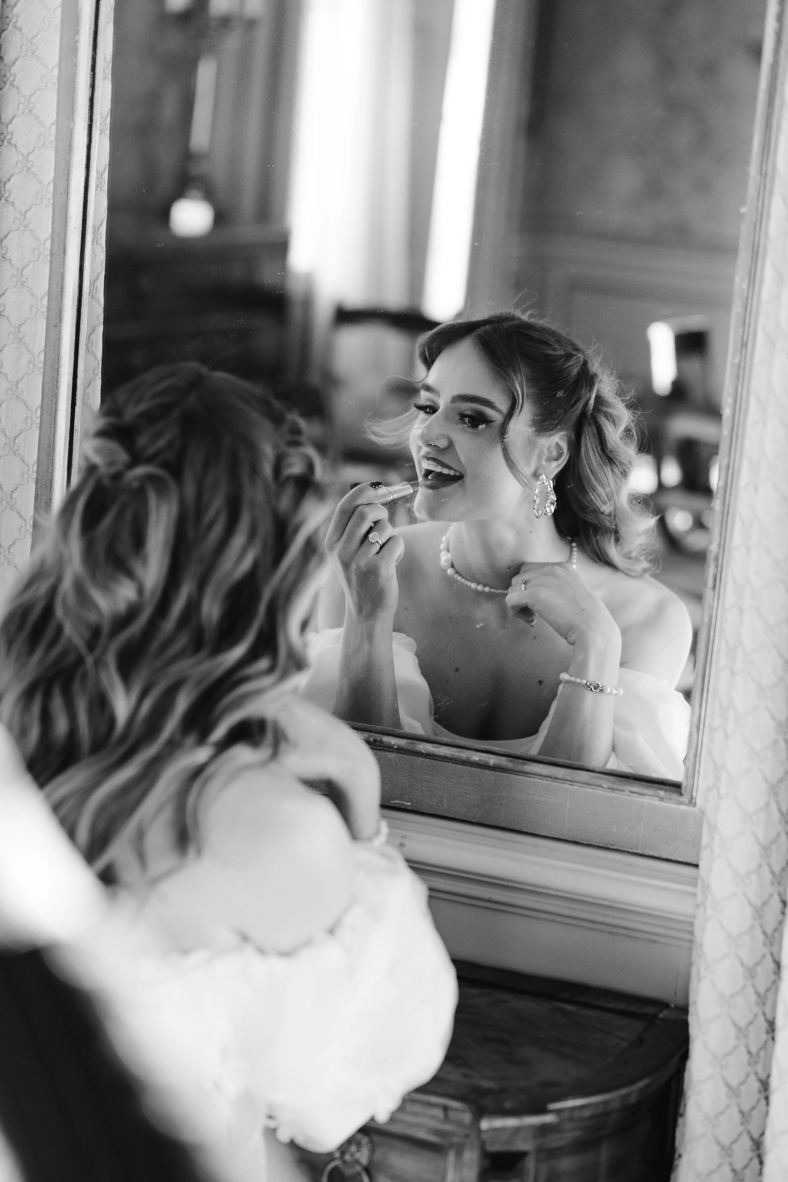 Een vrouw in formele kleding kijkt in de spiegel terwijl ze zich opmaakt, met een elegante uitstraling en sieraden die haar stijl aanvullen.