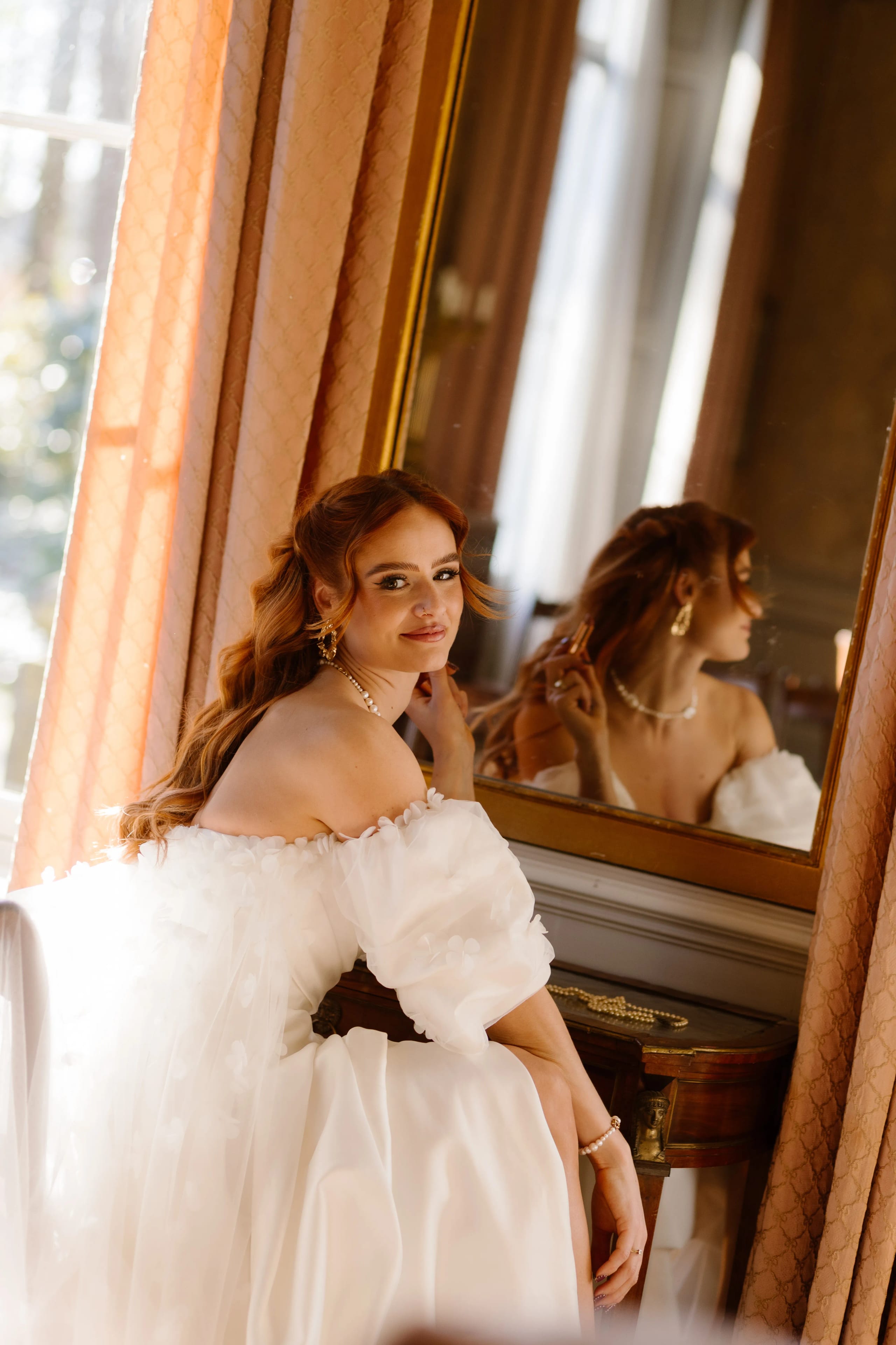 Een stijlvol aangeklede vrouw in een prachtige, witte trouwjurk zit ontspannen voor een spiegel. Haar lange, golvende haar en prachtige accessoires stralen elegantie uit, terwijl het zonlicht door het raam naar binnen valt en de warme sfeer van de kamer versterkt.