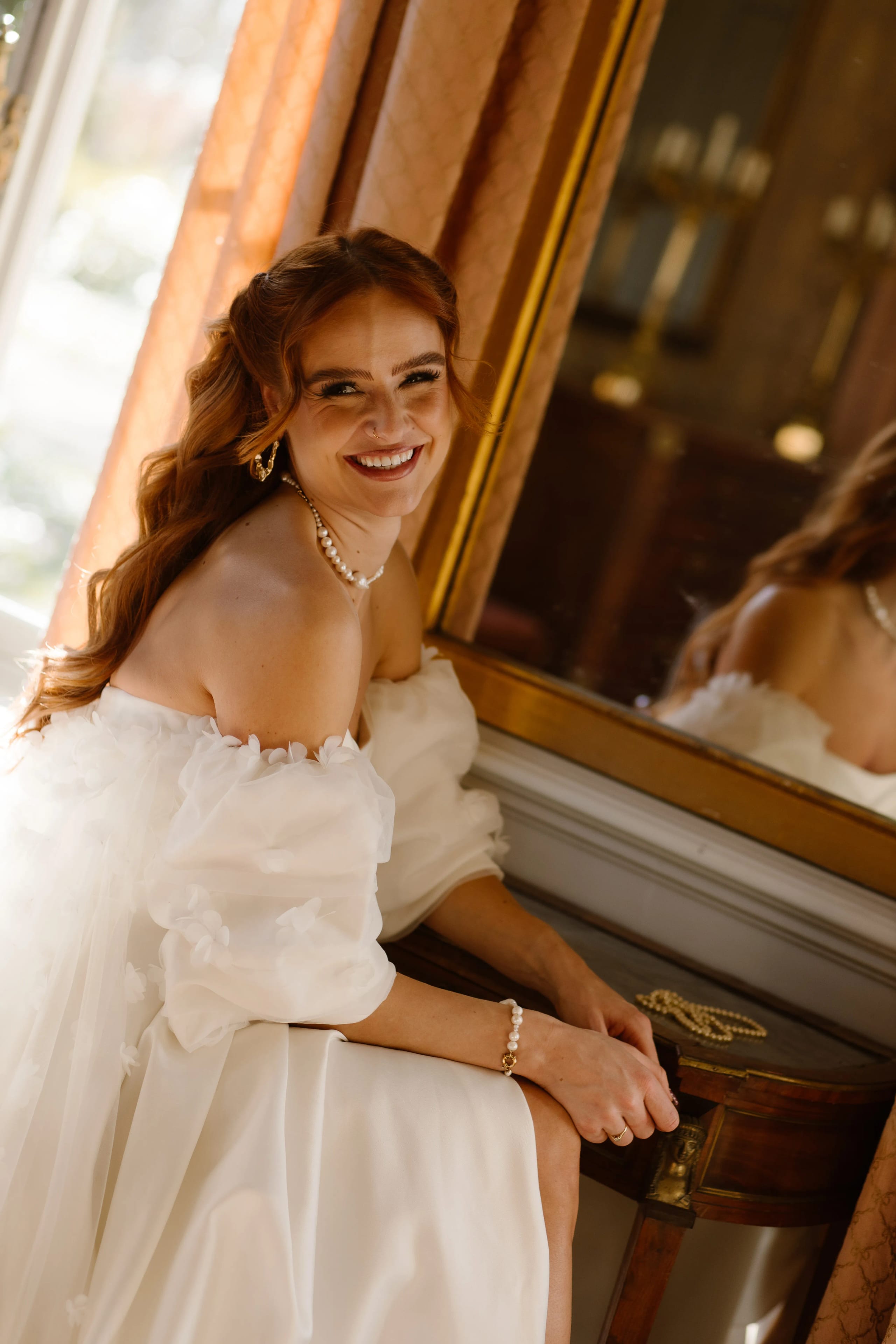 Een glimlachende vrouw in een prachtige witte jurk met pofmouwen zit ontspannen bij een spiegel, omringd door een elegante interieursetting met warme lichtinval. Haar glanzende rode haar en stijlvolle accessoires accentueren haar uitstraling van verfijning en schoonheid.