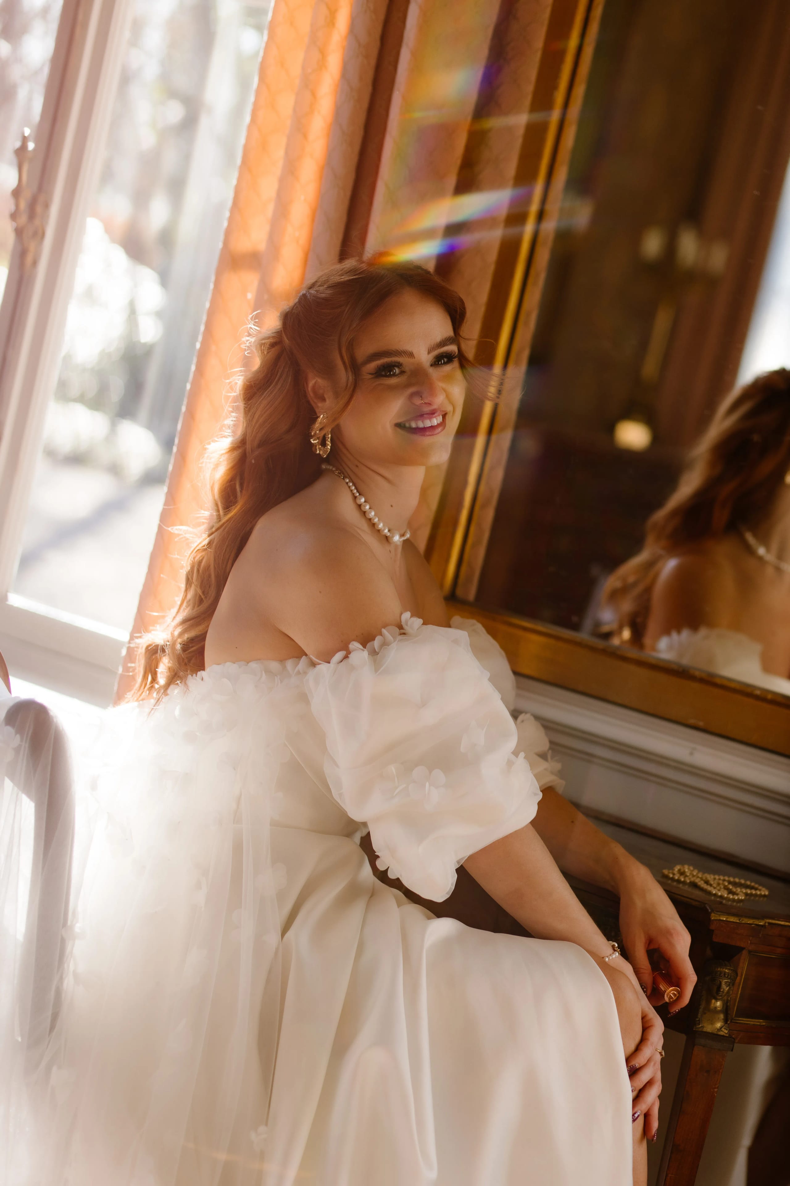 Een vrouw in een elegante witte trouwjurk met pofmouwen zit op een stoel bij een grote venster, met een glanzende spiegel op de achtergrond en een stralende glimlach die de romantische setting aanvult. Haar lange, golvende oren vallen op terwijl ze geniet van het zonlicht dat door het raam binnenvalt.
